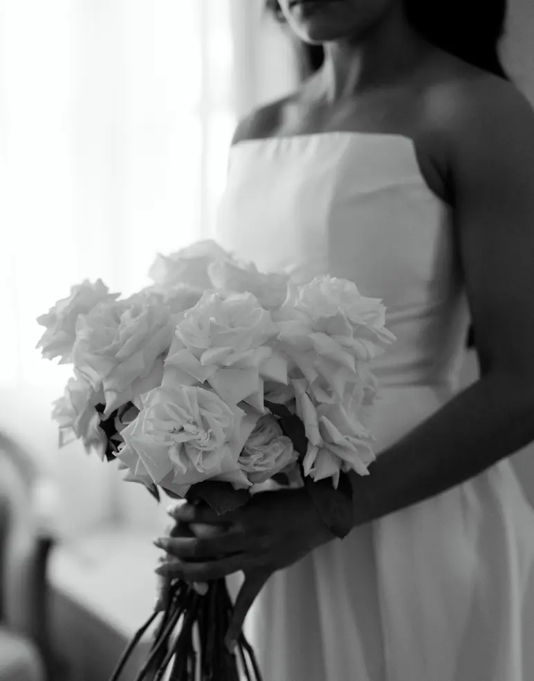 Black and white photo of a bride holding a large bouquet of white roses. The bride is wearing a strapless wedding dress and standing in a softly lit room, focusing on the flowers.