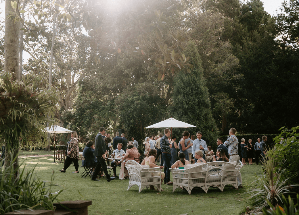 A group of people gathered outdoors in a lush, green garden setting. They are seated and standing around white wicker furniture and under white umbrellas. The scene is casual and social, with trees and plants surrounding the area under the soft sunlight.