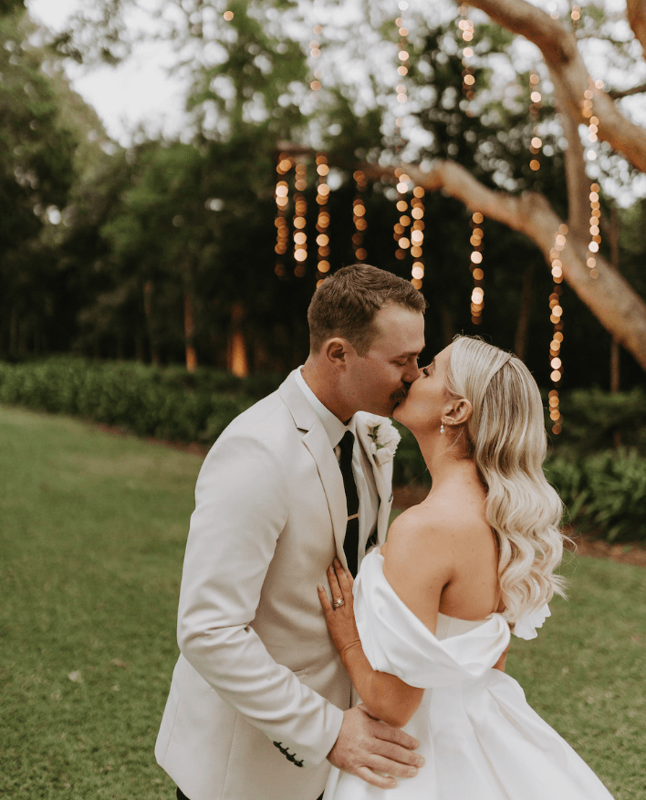 A couple dressed in formal attire shares a kiss outdoors during a wedding. Both wear light-colored outfits; the woman in a white gown with off-the-shoulder sleeves and the man in a beige suit. Strings of lights hang from a tree in the background, enhancing the romantic ambiance.