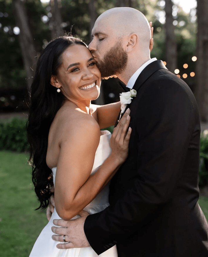 A bride and groom share an intimate moment outdoors. The bride, wearing a strapless white dress, smiles radiantly as the groom, in a black suit, gently kisses her forehead. They are surrounded by lush greenery and twinkling string lights in the background.