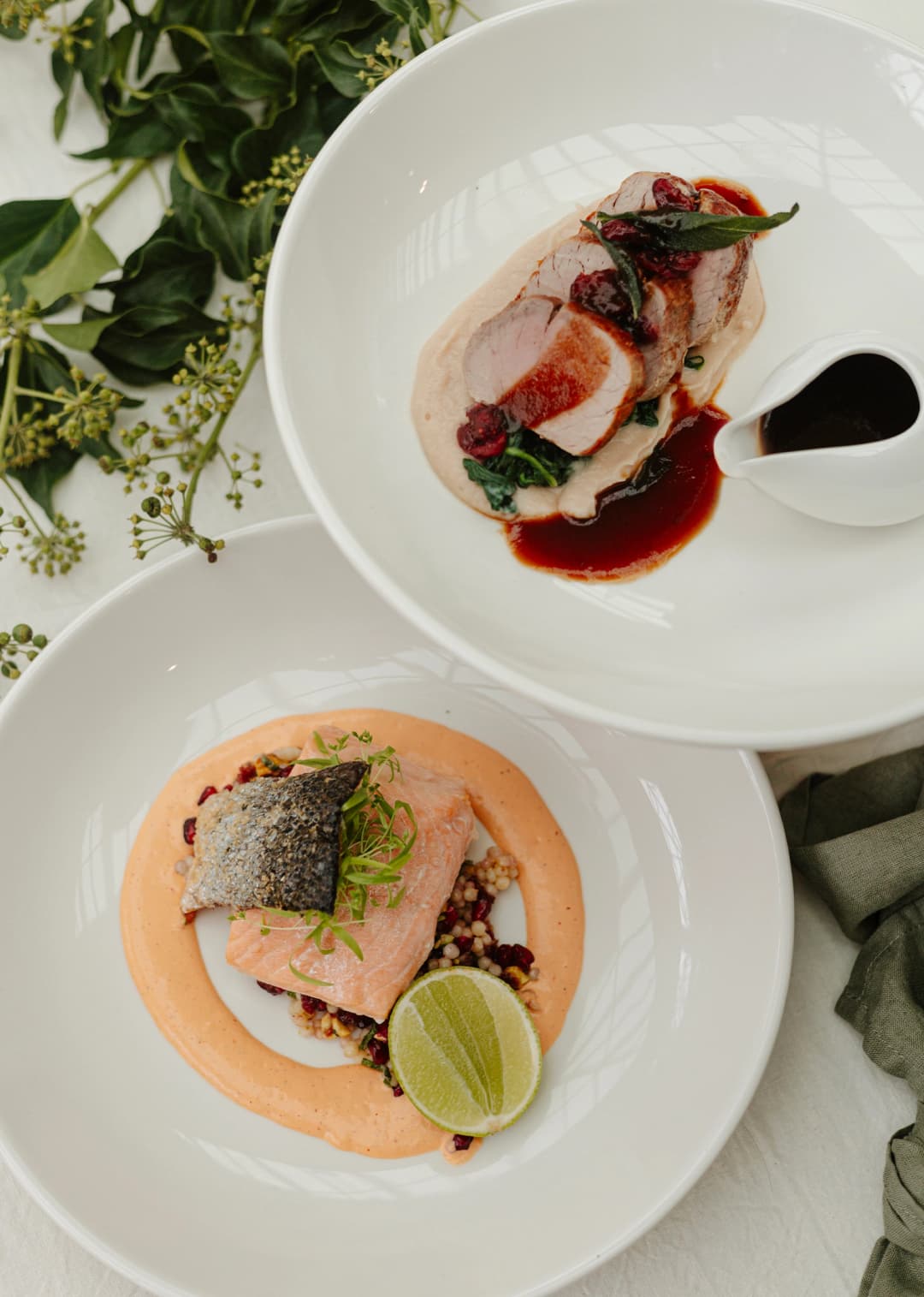 Two white plates of gourmet food are elegantly presented. The top plate features meat slices with greens, sauce, and berries. The bottom plate showcases a piece of fish with crispy skin, resting on a bed of grains, garnished with sauce, scallions, and a lime wedge.