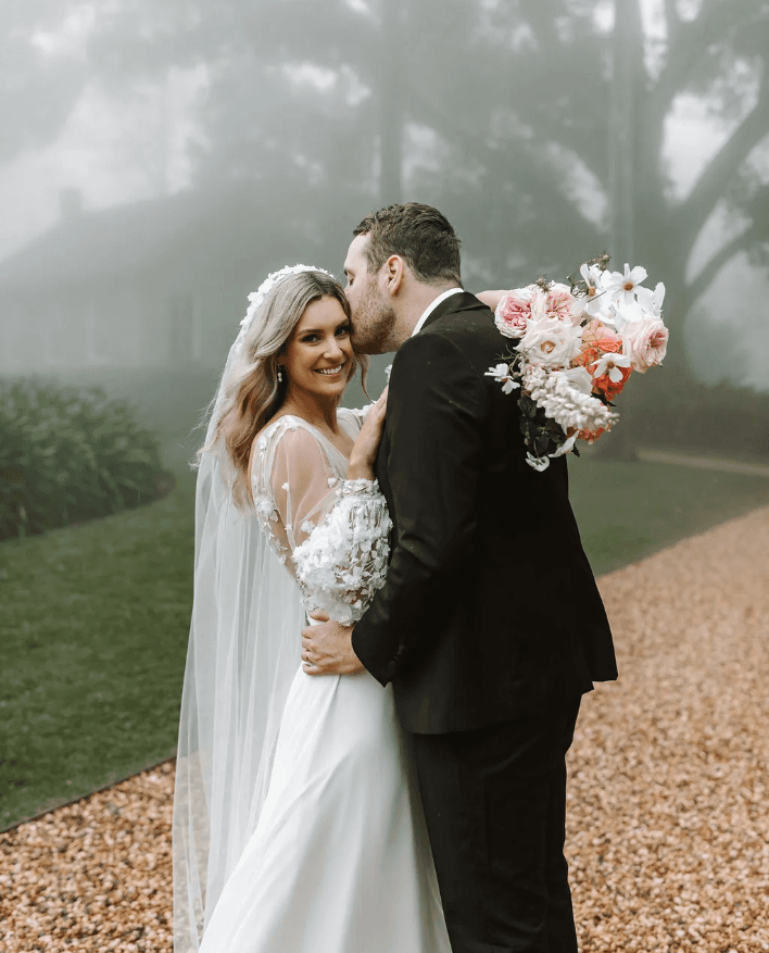 A bride and groom share a tender moment on a foggy day. The groom kisses the bride on the temple while she smiles at the camera. The bride wears a white dress and veil, holding a bouquet of pink and white flowers. The groom is dressed in a black suit.