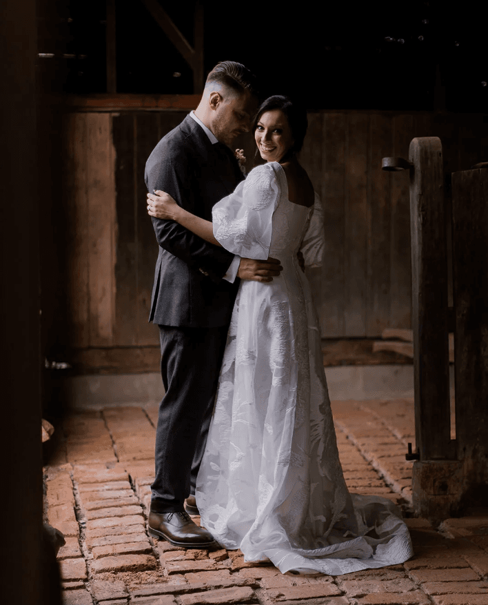 A bride and groom share an intimate moment in a rustic setting, standing on a red brick floor. The groom is in a dark suit, and the bride wears a white dress with lace details. They embrace, gazing at each other with warm smiles, creating a tender and romantic scene.