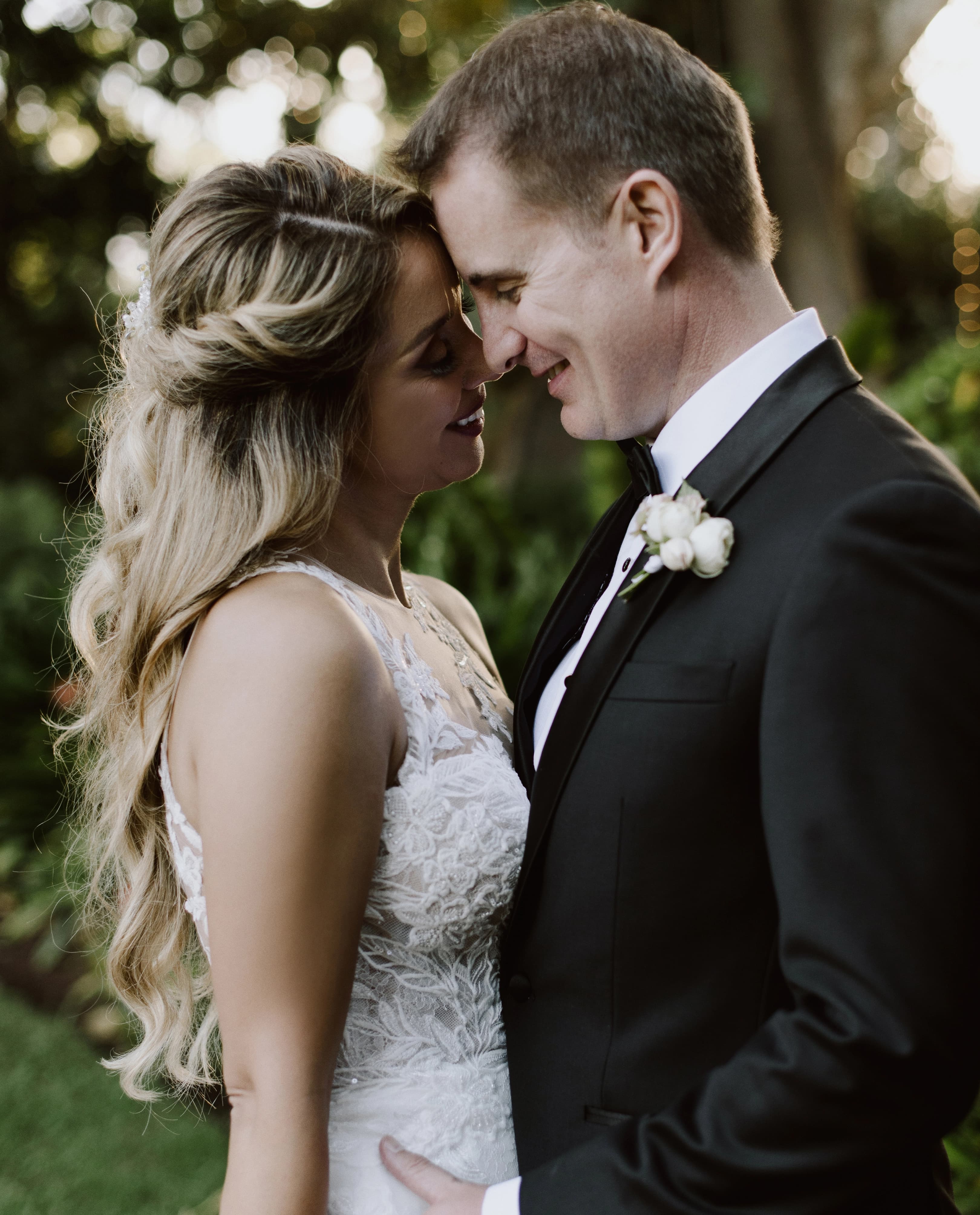 A bride and groom share an intimate moment outdoors, touching foreheads and smiling at each other. The bride has long blonde hair styled in loose curls and wears a white lace dress. The groom wears a classic black suit with a white boutonniere on his lapel.