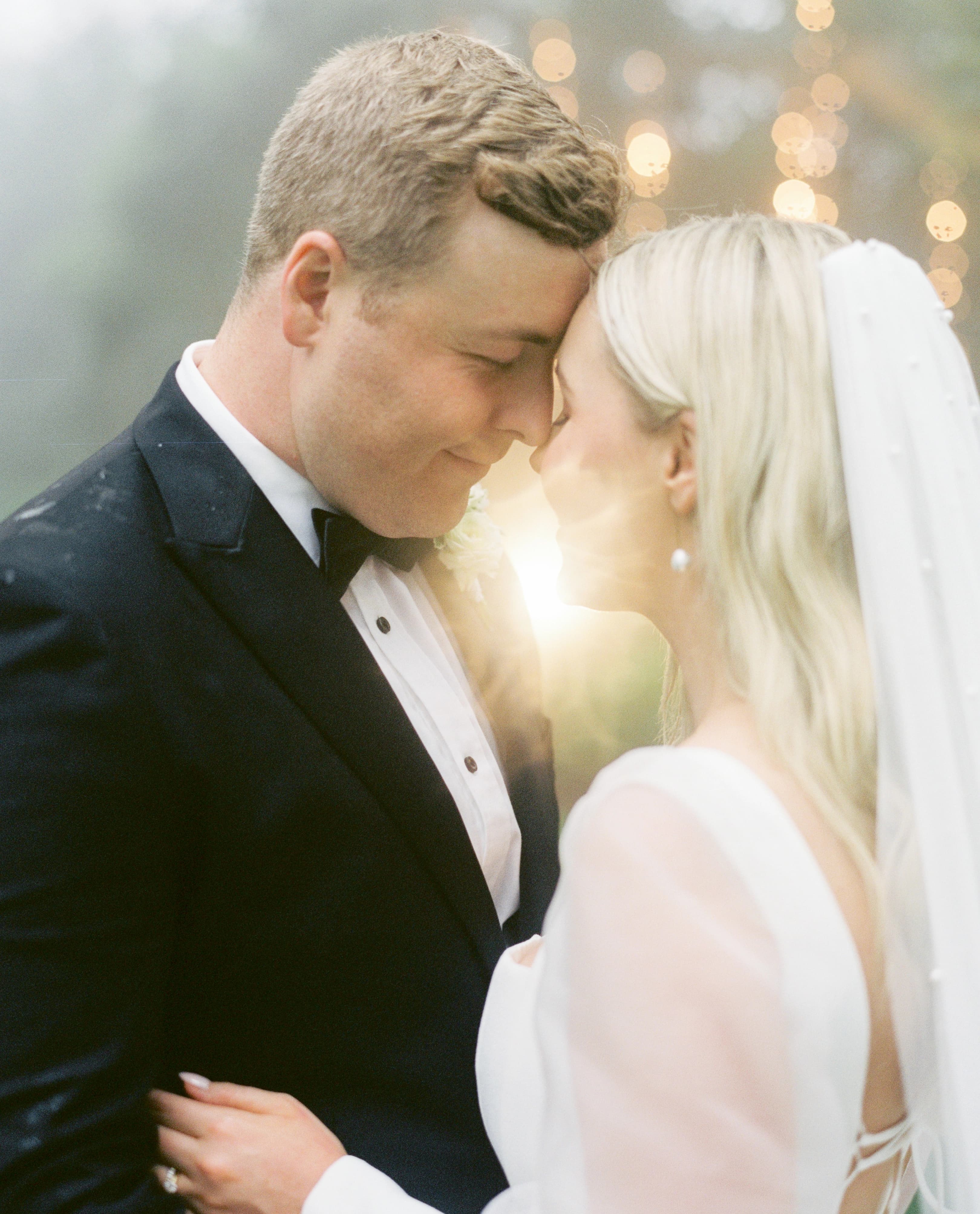 A newlywed couple gently touch foreheads, sharing an intimate moment. The groom, in a black suit and bow tie, faces the bride, who is dressed in a white wedding gown with a veil. Warm, soft lighting in the background creates a romantic ambiance.