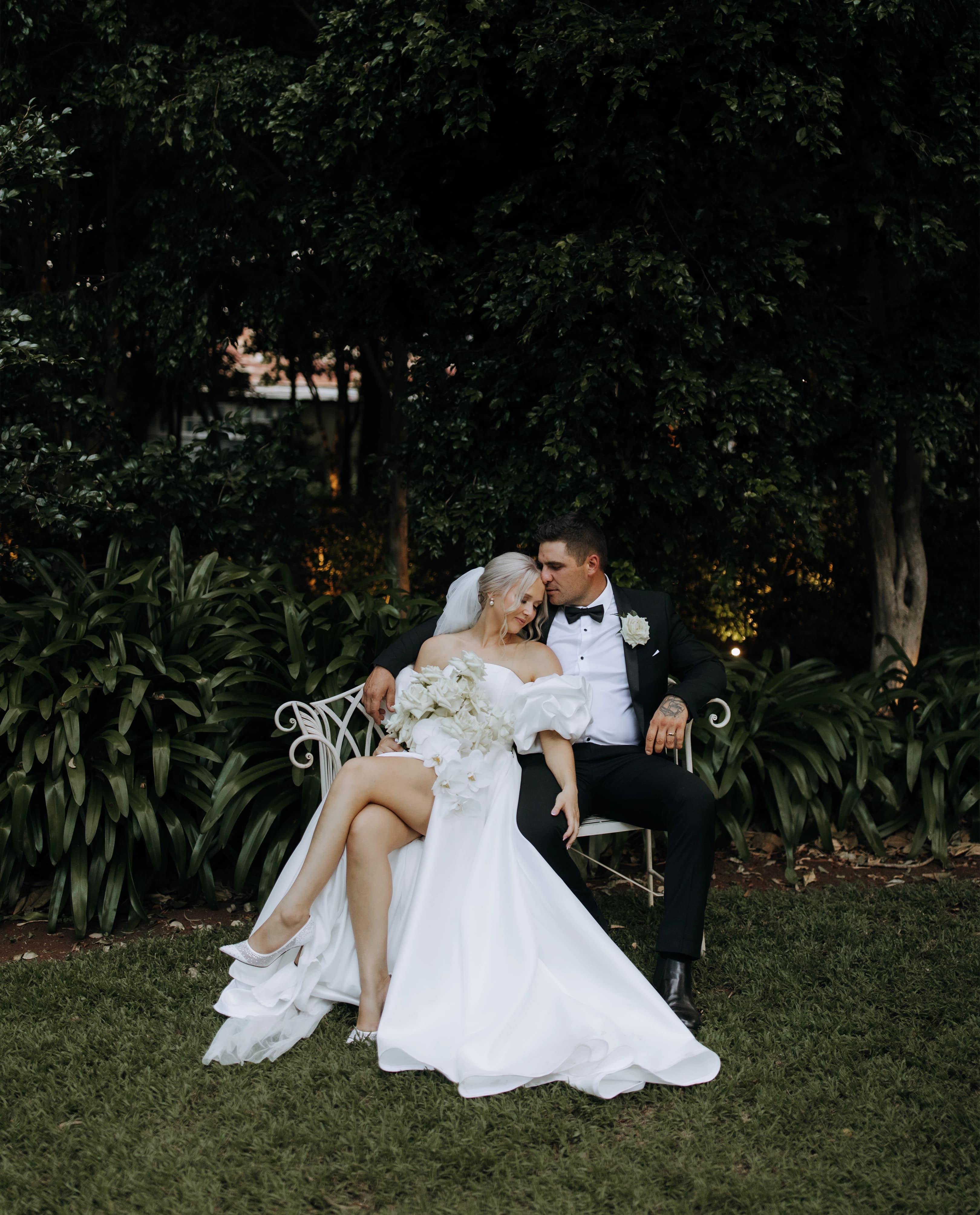 A bride and groom sit on a white bench in a garden. The bride, in a white dress holding a bouquet, has her legs draped over the groom's lap. The groom, wearing a black tuxedo, and bride gaze at each other lovingly as they pose in front of lush greenery.