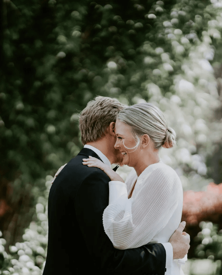 A couple embraces outdoors with a backdrop of greenery. The man, in a black suit, has his back to the camera, while the woman, in a white dress, smiles and rests her chin on his shoulder. The scene is soft and serene with sunlight filtering through the foliage.