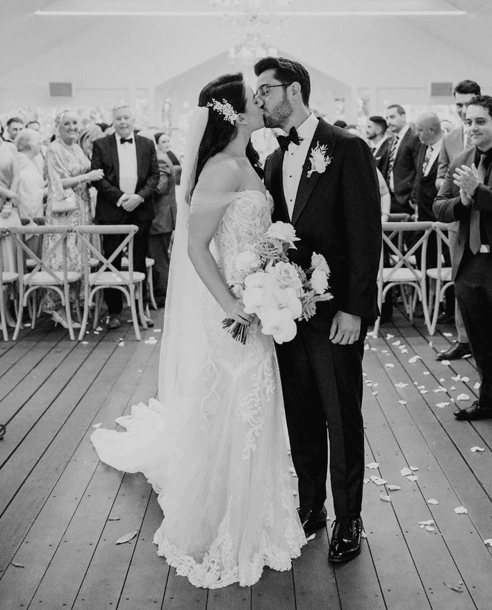 A bride and groom share a kiss at their wedding ceremony. The bride is wearing an off-the-shoulder lace gown and holding a bouquet of flowers, while the groom is in a classic tuxedo. Guests stand and applaud in the background, and rose petals are scattered on the floor.