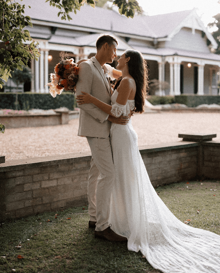 A newlywed couple is embracing outdoors. The groom, in a light beige suit, and the bride, in a white, off-shoulder wedding dress with a long train, gaze into each other's eyes. They are standing on a lush lawn with a charming house in the background.
