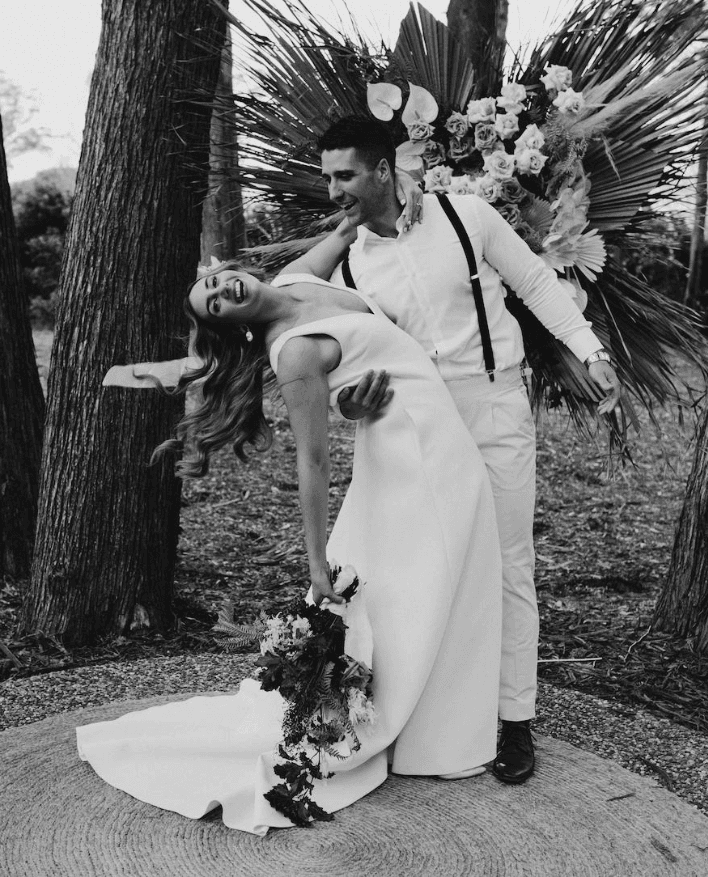 A smiling couple, dressed in wedding attire, pose playfully in a forest. The groom, wearing suspenders, dips the bride, who holds a bouquet and wears a flowy dress. Behind them is a backdrop of large flowers and palm fronds. The image is in black and white.