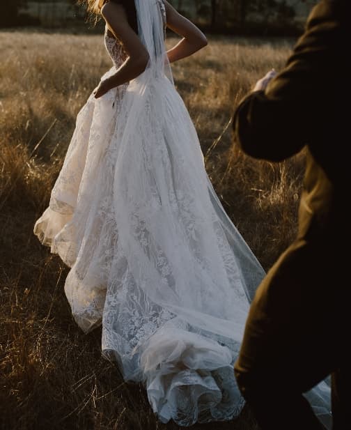 Bride in a flowing white lace wedding gown walks through a field of tall grass at sunset, with a person in dark clothing slightly blurred in the foreground. The gown features intricate detailing and a long, elegant train trailing behind her.