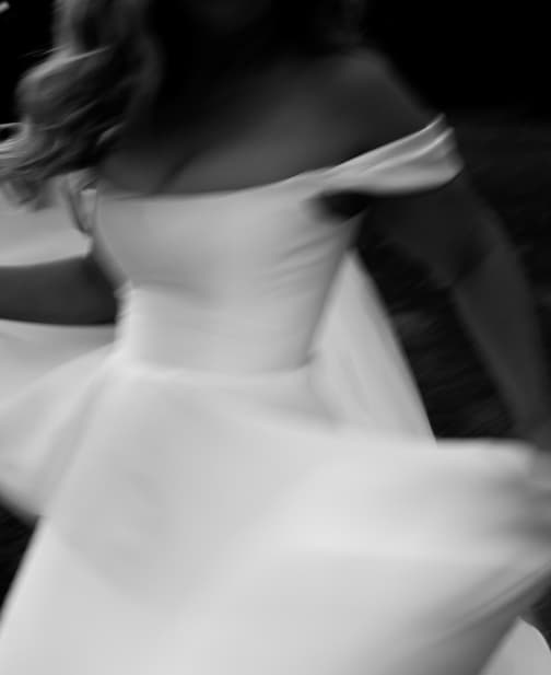 A black and white blurred image of a person in an off-the-shoulder dress spinning or moving quickly, creating a sense of motion. The focus is on the dress, which appears to be formal and flowing. The person's face is not clearly visible.