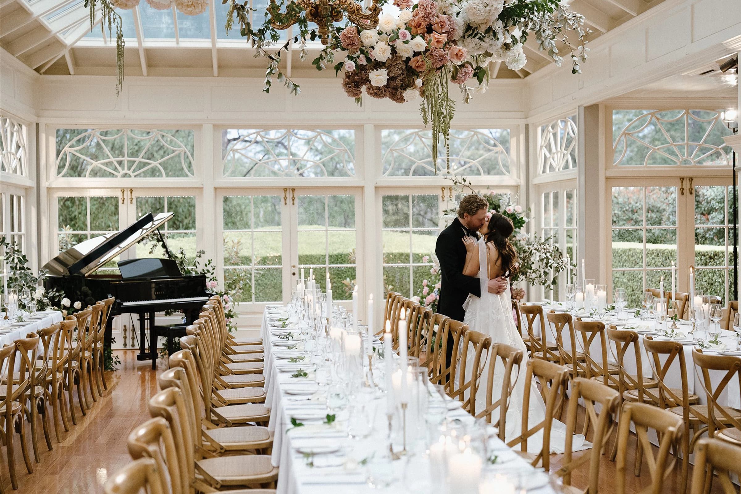 A couple in wedding attire share a kiss in an elegant, bright reception hall. The room is decorated with a hanging floral arrangement and long dining tables set for guests. Large windows overlook a lush garden, and a grand piano is positioned in one corner.