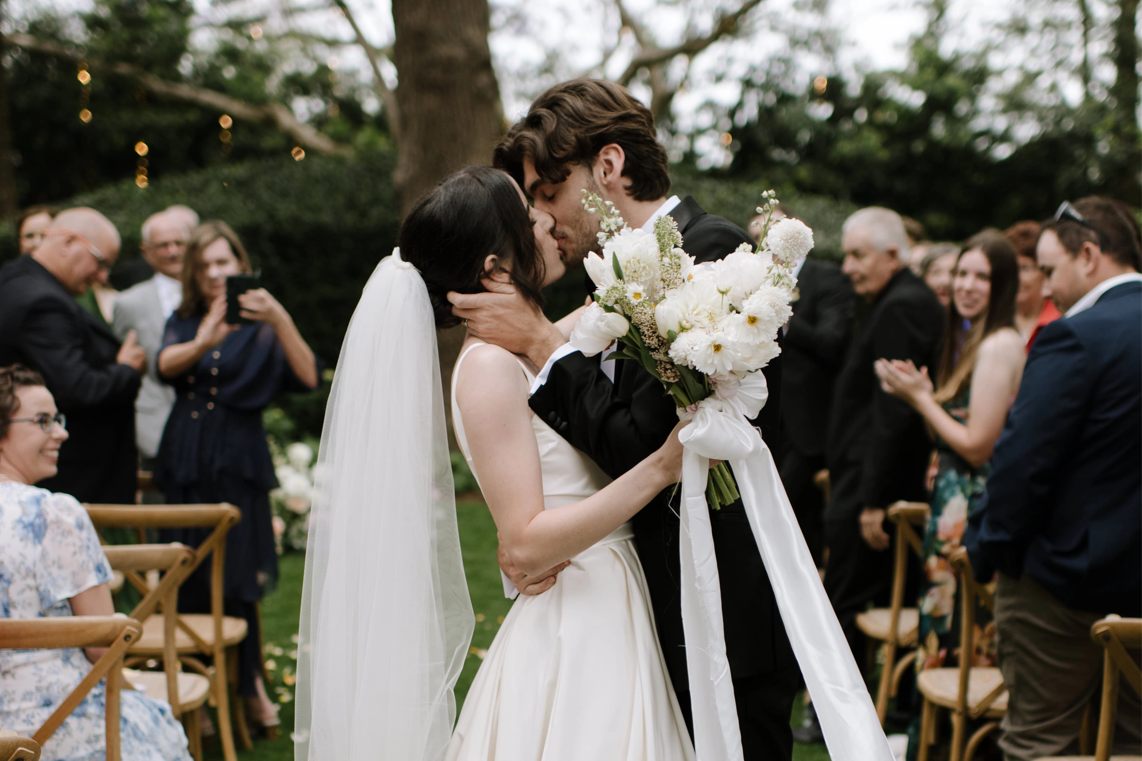 A bride and groom share a kiss at an outdoor wedding ceremony. The bride, in a white gown and veil, holds a bouquet of white flowers. The groom is dressed in a dark suit. Guests are seated and standing around them, clapping and taking photos. Trees are visible in the background.