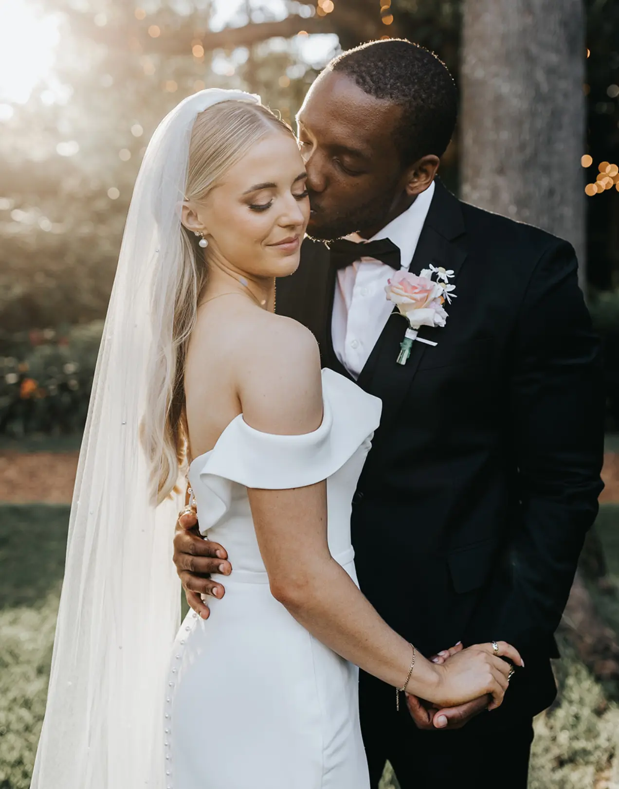 A bride in a white dress and veil smiles with her eyes closed as her groom, wearing a black suit with a pink rose boutonnière, gently kisses her forehead. They stand outside surrounded by trees and sunlight, holding hands and embracing closely.