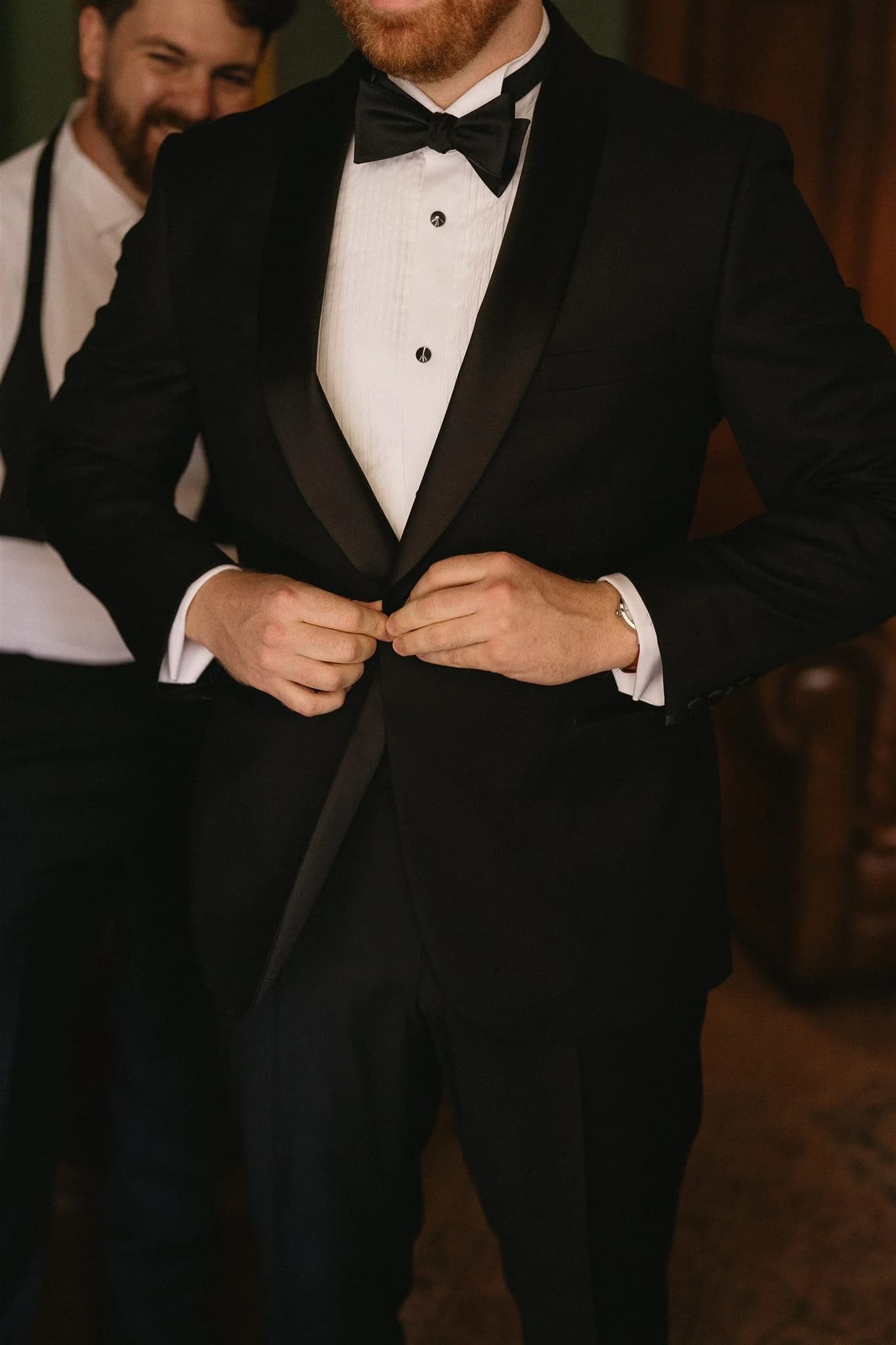 A man wearing a black tuxedo with a white dress shirt adjusts his jacket. He has ginger hair and a beard. Another man in a tuxedo stands behind him, smiling. The background is a warmly lit room with wooden furniture.