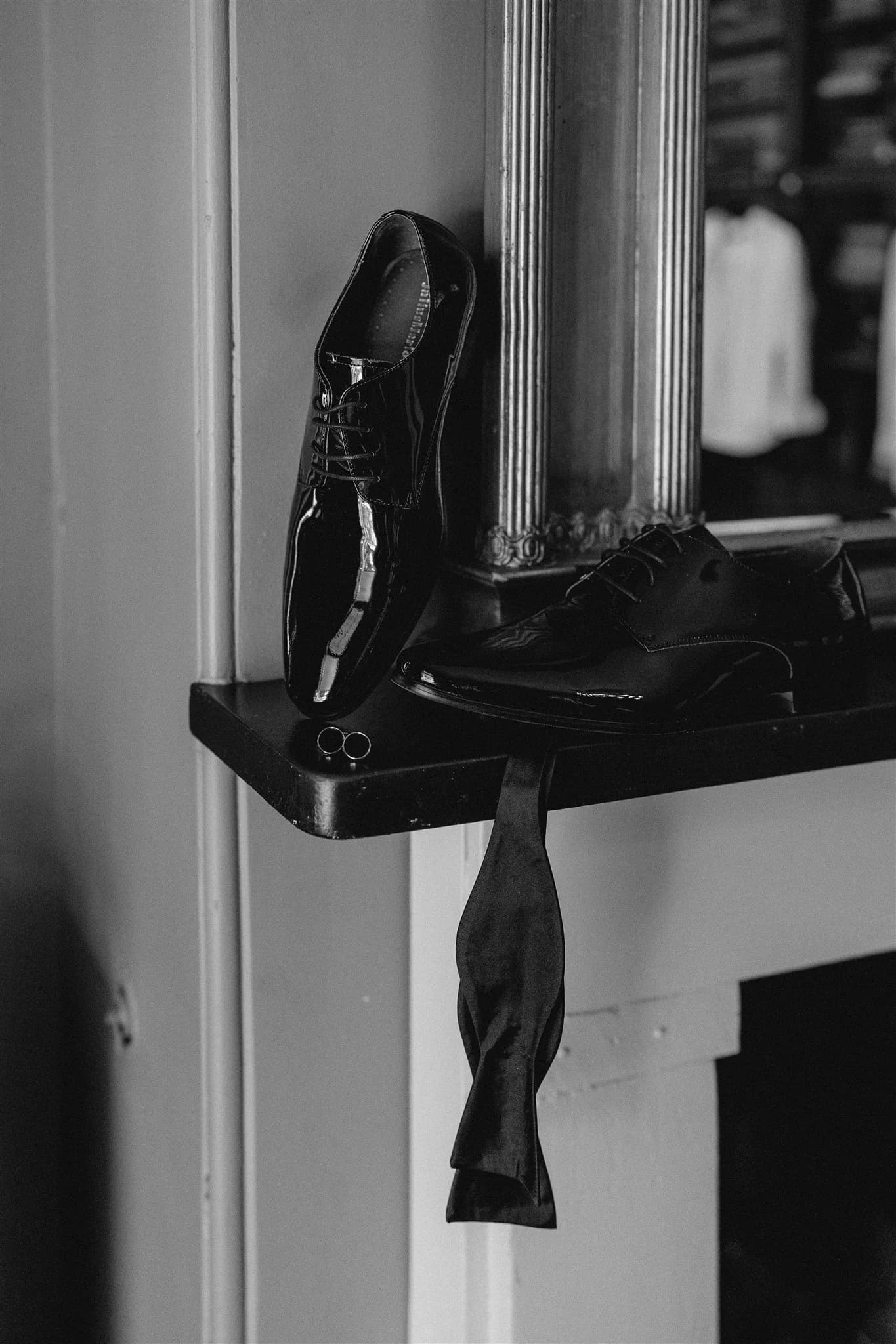 Black and white image of a pair of shiny dress shoes placed on a shelf next to a pillar. A black bow tie hangs loosely from the shelf. The background includes a blurred view of a clothing rack with hanging shirts.