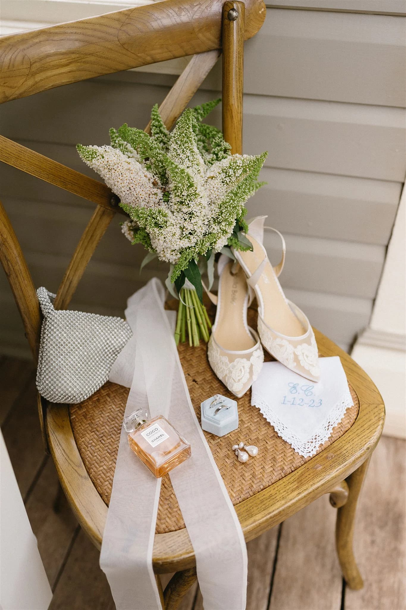 A wooden chair holds a bouquet of white flowers, a pair of lace bridal shoes, a silver clutch, a perfume bottle, earrings, and a handkerchief with "8-6-1123" embroidered in blue. A white ribbon is draped across the chair, creating a serene wedding preparation scene.