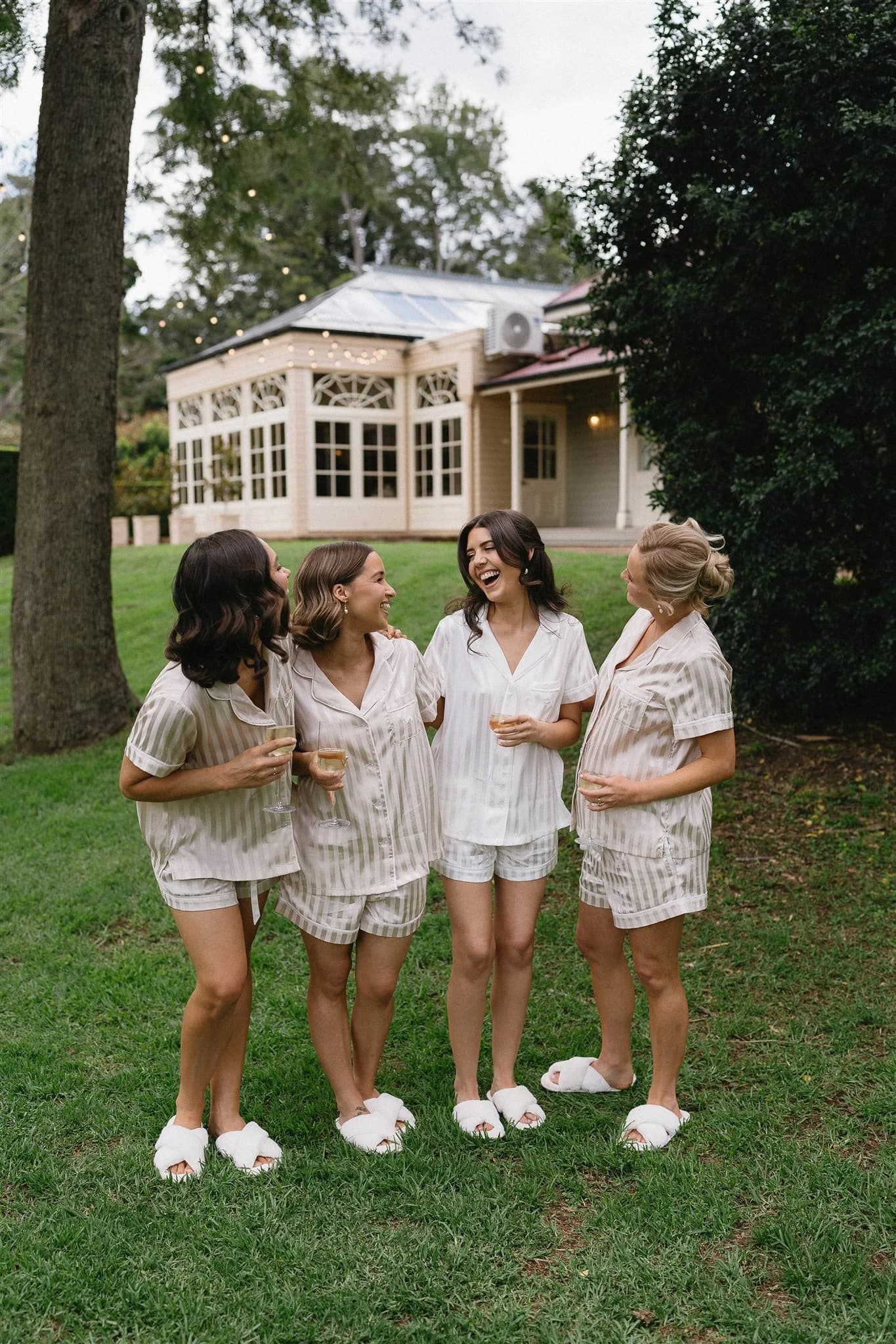 Four women wearing matching pajamas and slippers stand on a lawn, laughing and chatting. They are in front of a large building with a glass-paned facade and surrounded by trees and greenery.