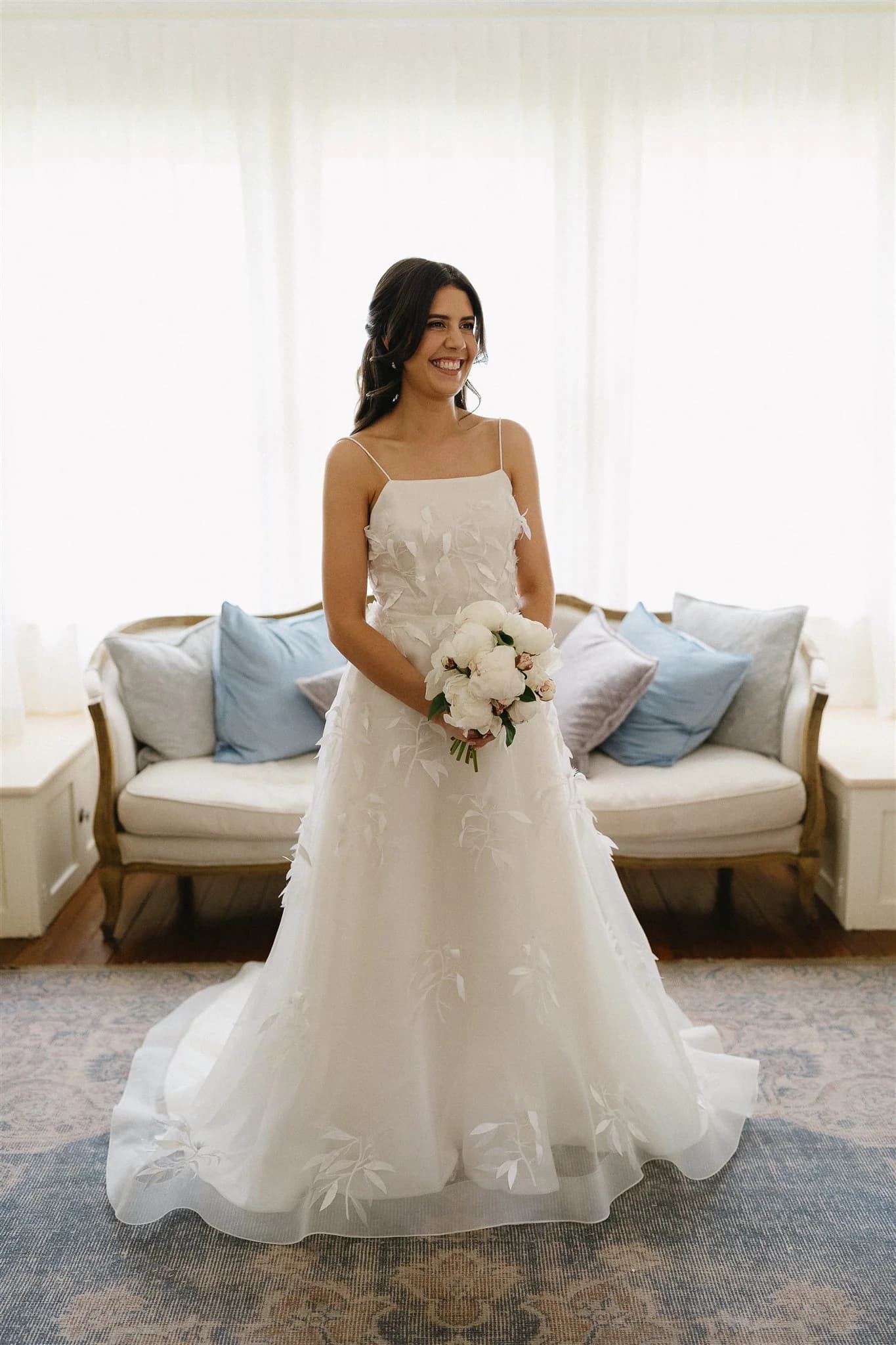 A bride stands indoors in front of a white curtain, smiling and holding a bouquet of white flowers. She wears a white, sleeveless wedding gown with floral appliqués. Behind her is a white sofa adorned with blue and gray pillows.