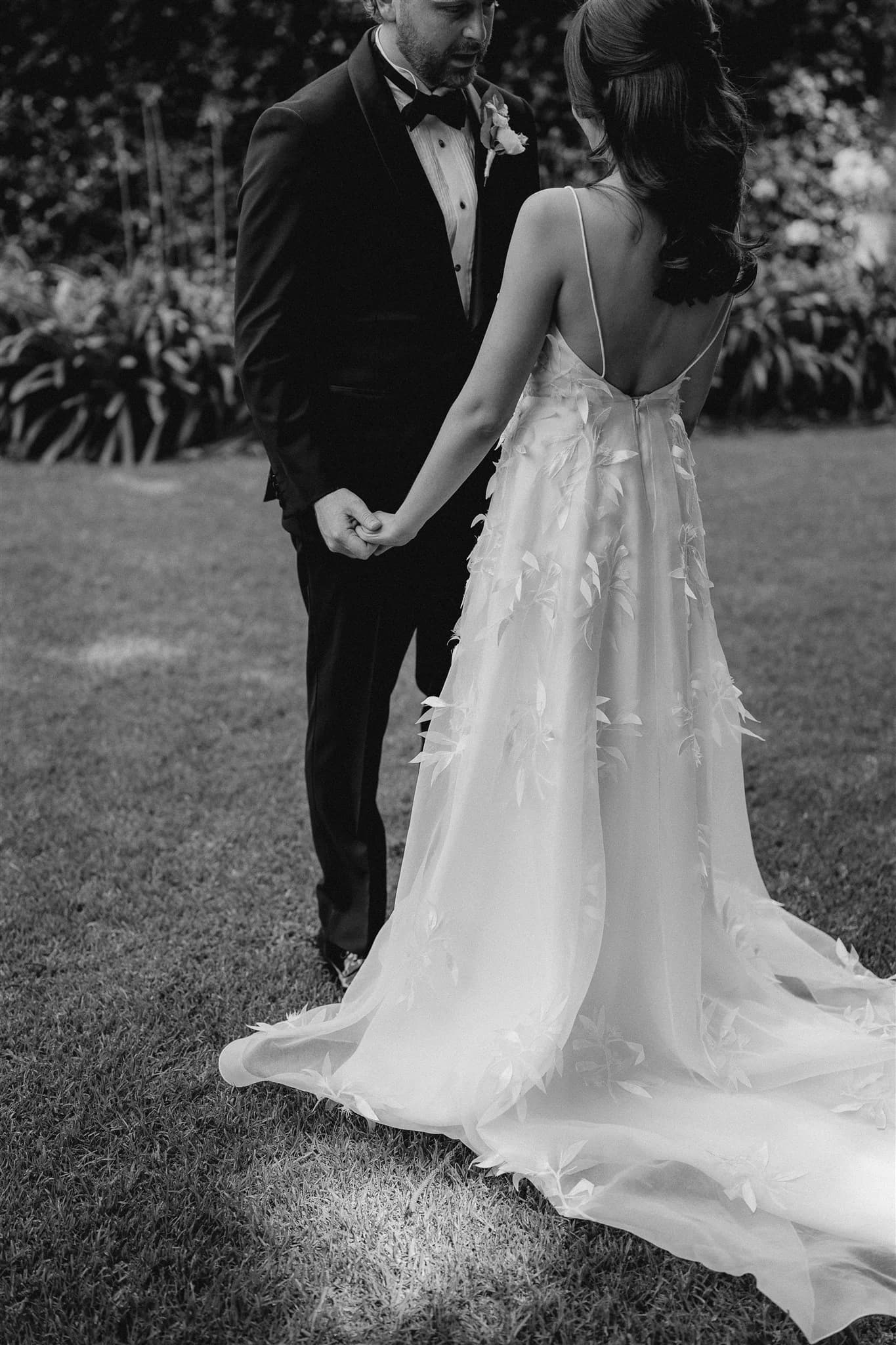 A black-and-white photo of a couple on their wedding day. The bride, in a long, flowing sleeveless dress with floral embellishments, holds hands with the groom, who is dressed in a dark suit and bowtie. They stand on a grassy lawn with greenery in the background.