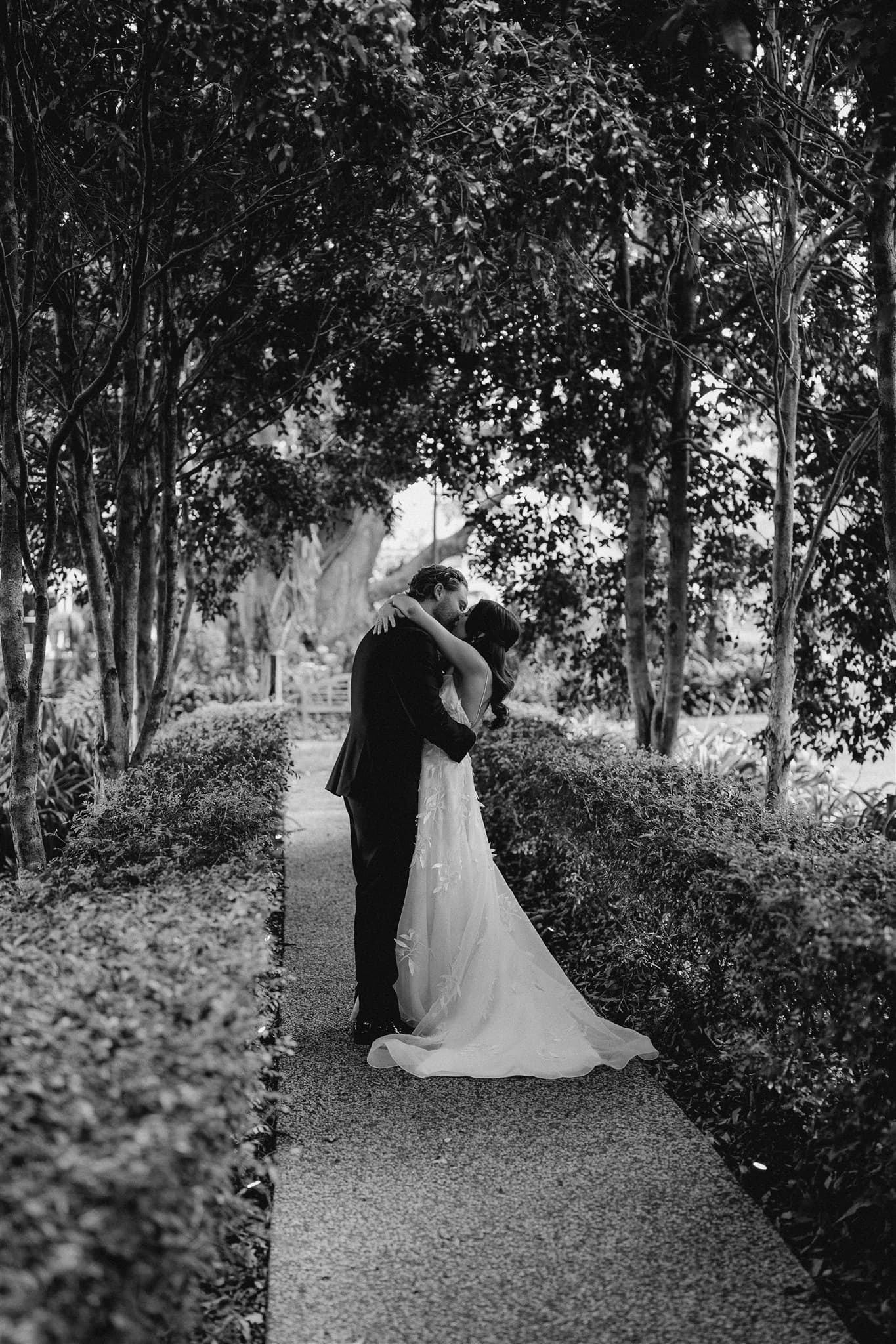 Black and white image of a couple embracing on a narrow, tree-lined path. The bride is in a flowing, strapless gown, and the groom is in a dark suit. They are surrounded by greenery and the overhead trees create a natural archway above them.