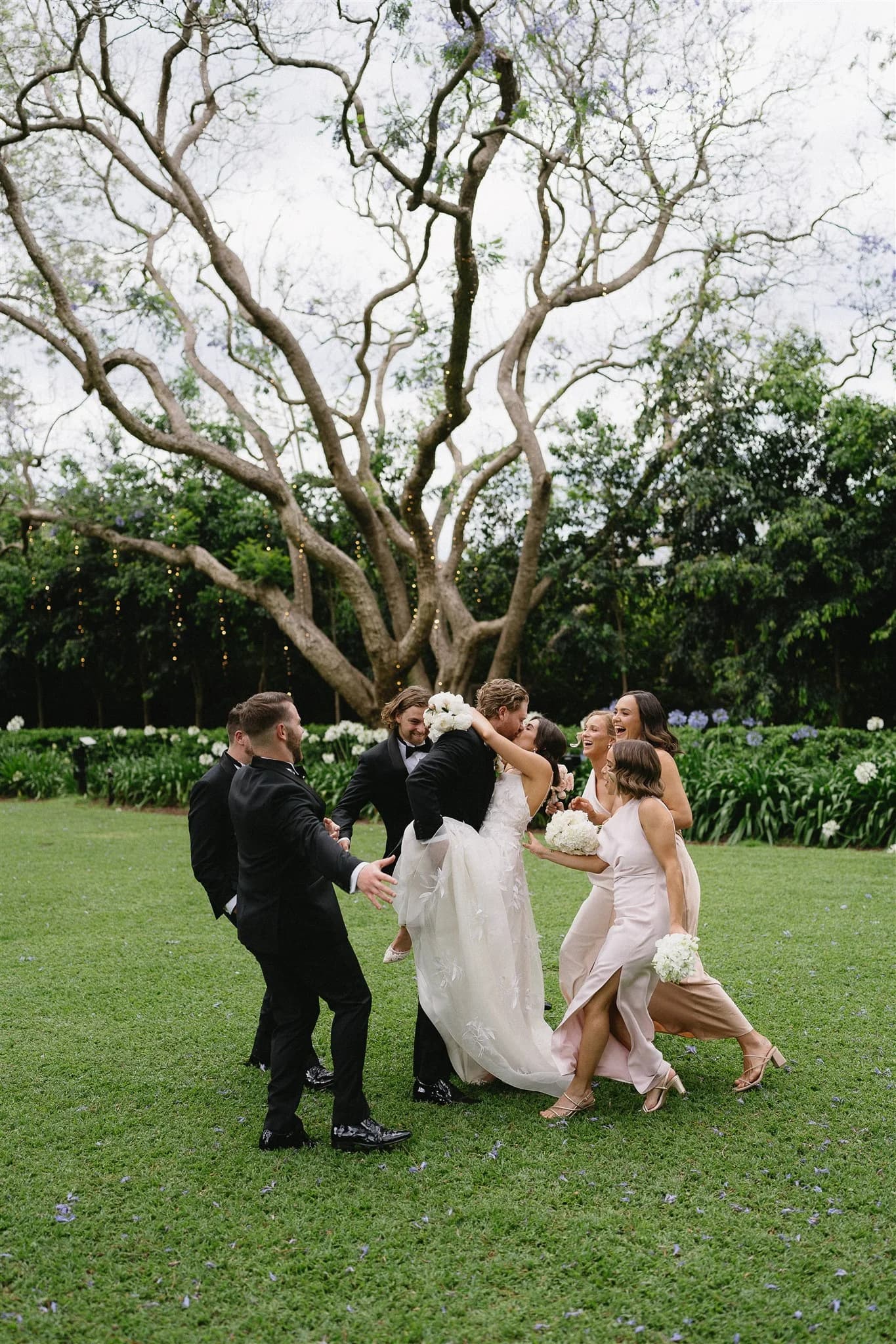 A joyful wedding scene with a bride in a white gown being lifted and embraced by a group of people, including bridesmaids in light pink dresses and groomsmen in black suits. They are standing on a lush green lawn, with a large tree and greenery in the background.