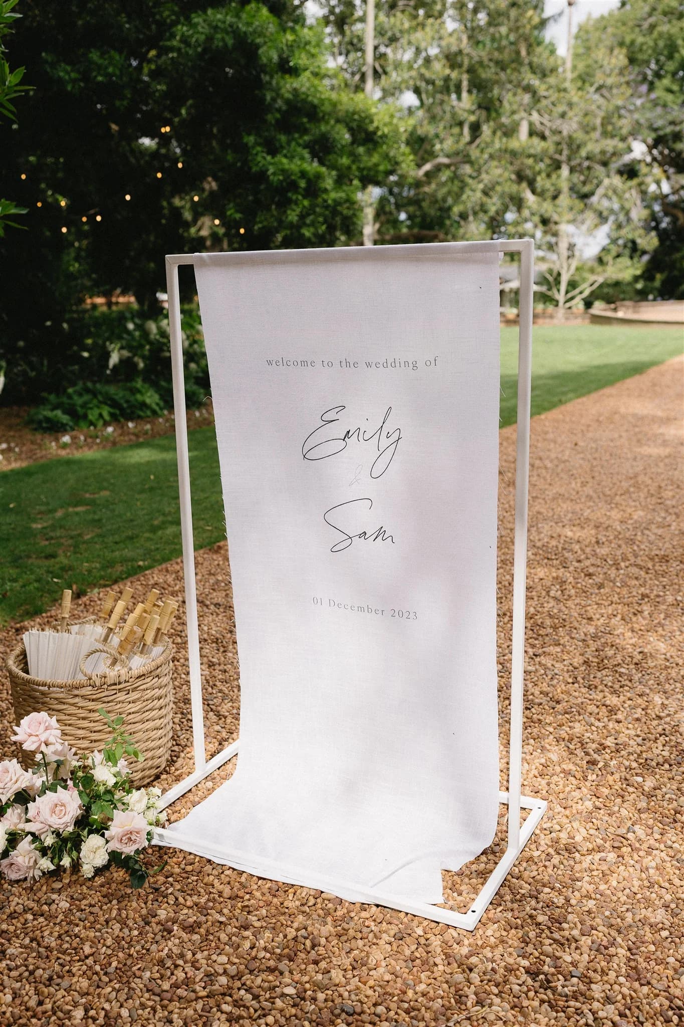 A white wedding sign framed in metal stands on a gravel path, surrounded by greenery. The sign reads "Welcome to the wedding of Emily & Sam, 01 December 2023." A basket filled with rolled papers and a small bouquet of flowers are placed nearby.