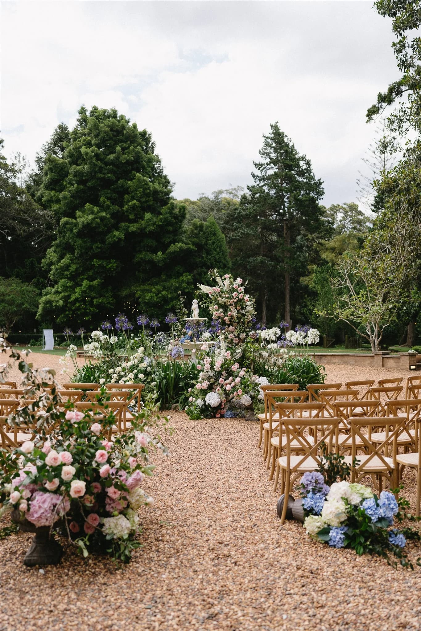 Outdoor wedding ceremony setup in a garden. Wooden chairs are arranged in rows on a gravel path, adorned with floral arrangements of pink, white, and blue flowers. An archway of flowers frames the altar in the distance, surrounded by lush greenery and trees.