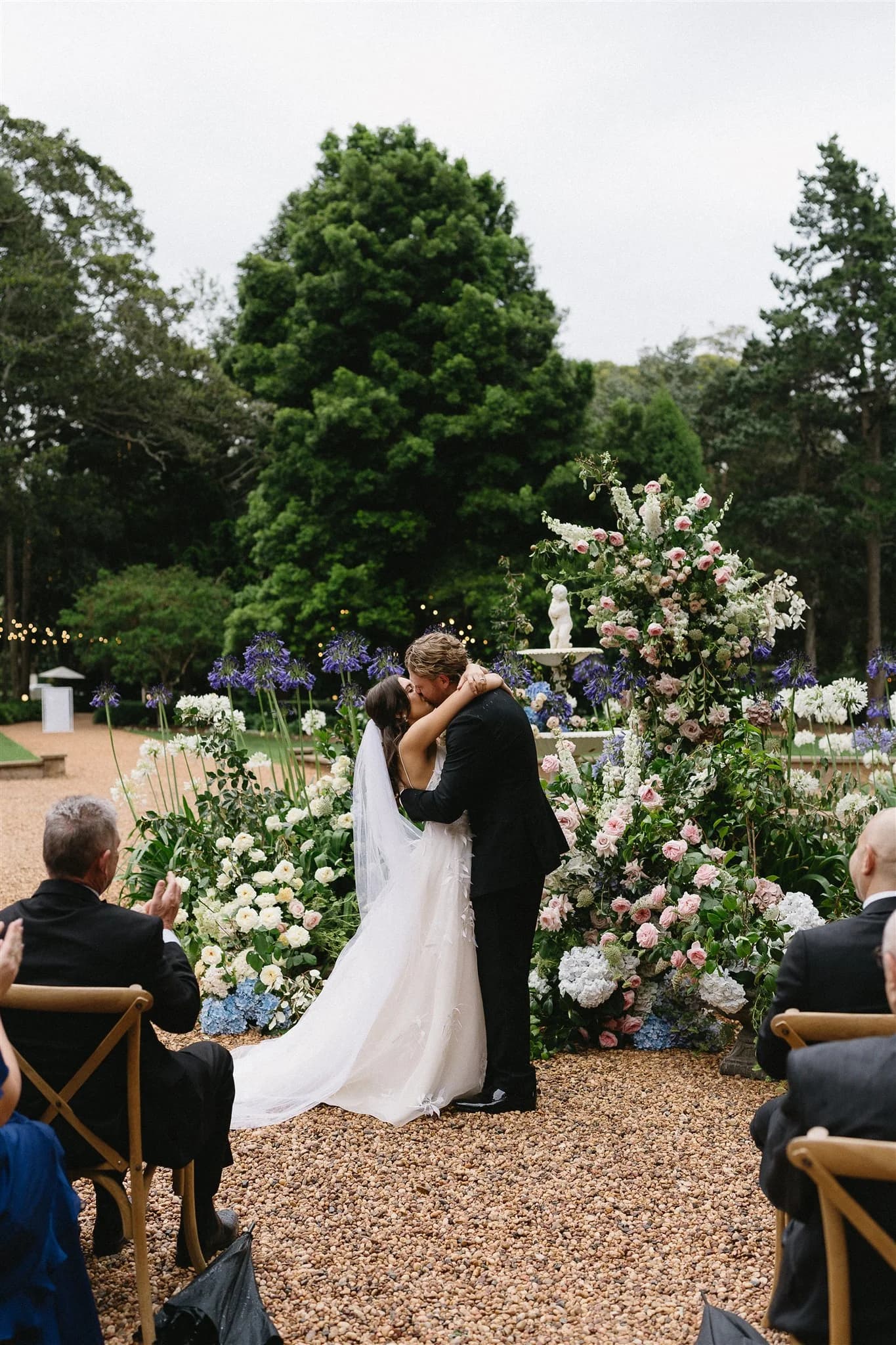 A newlywed couple shares a kiss at an outdoor wedding ceremony. They stand in front of a lush, flower-adorned altar with vibrant greenery and floral arrangements. Guests sit on wooden chairs, some clapping, while trees and a gravel pathway complete the serene setting.