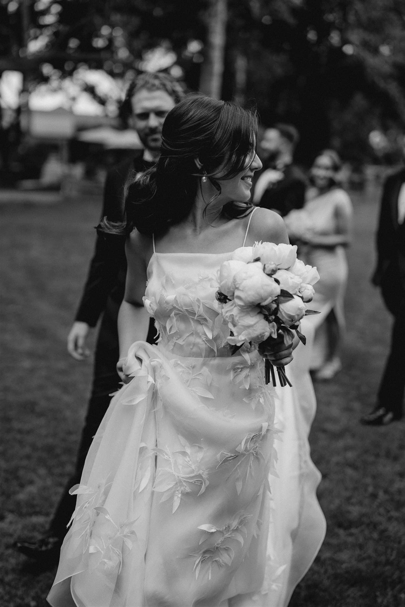 Black and white photo of a smiling bride holding a bouquet while walking outdoors with a group of people. The bride is wearing an elegant dress adorned with floral appliqués and is glancing to her left. Other guests are visible in the background.