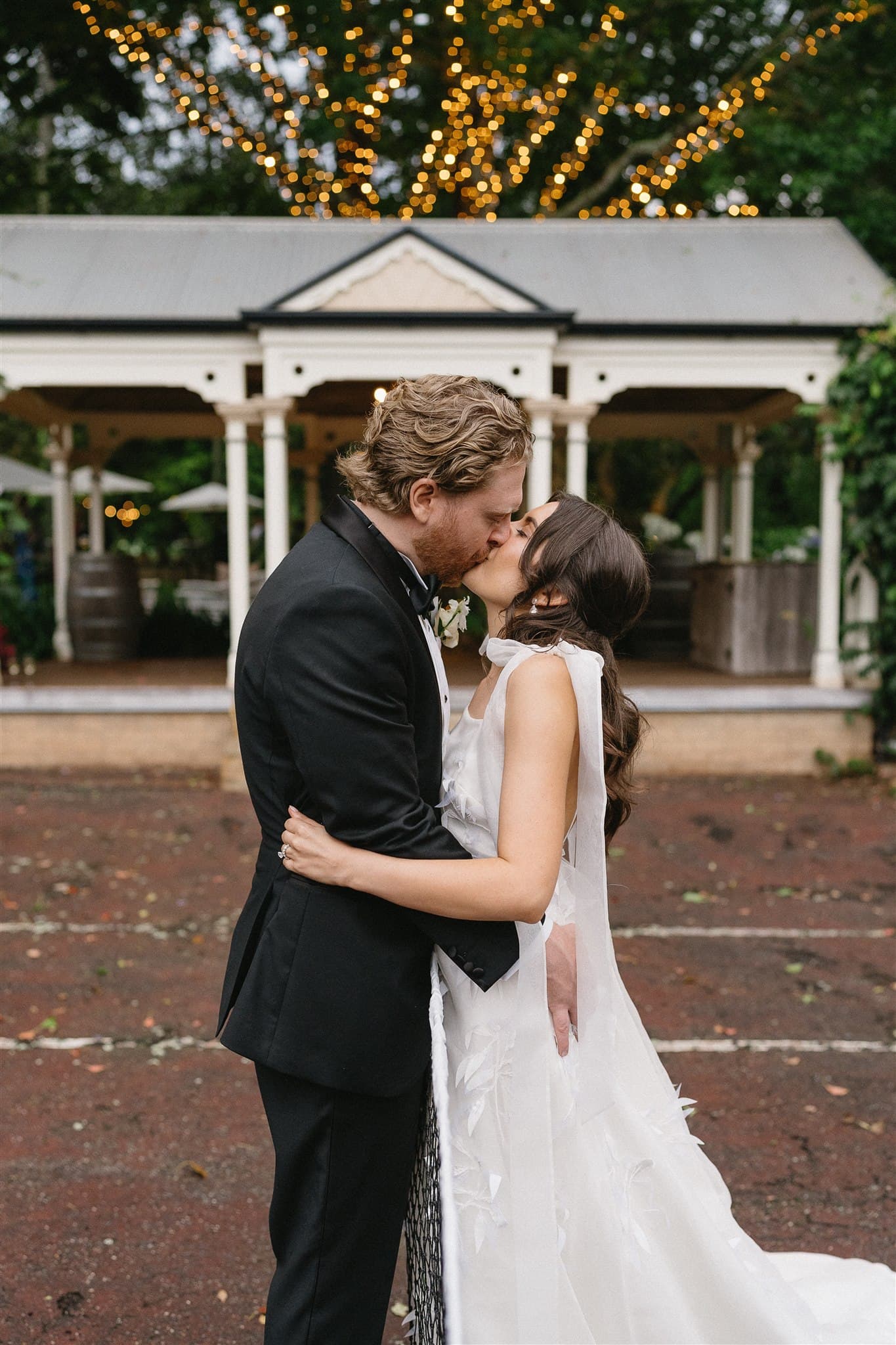 A couple in formal attire shares a kiss outdoors. The man, in a black suit, embraces the woman dressed in a flowing white gown. They stand on a red brick surface with a gazebo adorned with string lights in the background. It appears to be a wedding setting.