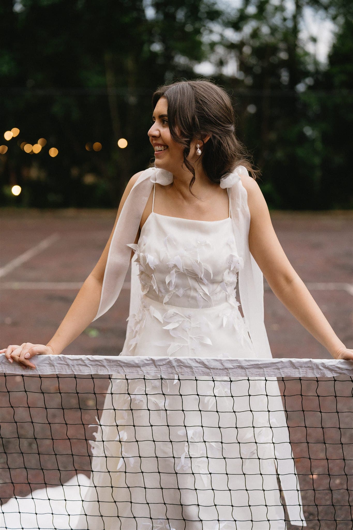 A young woman in a white dress with decorative feathers stands smiling, leaning on a tennis net. The background is an outdoor tennis court surrounded by trees and softly glowing string lights. The scene exudes a whimsical, joyful atmosphere.