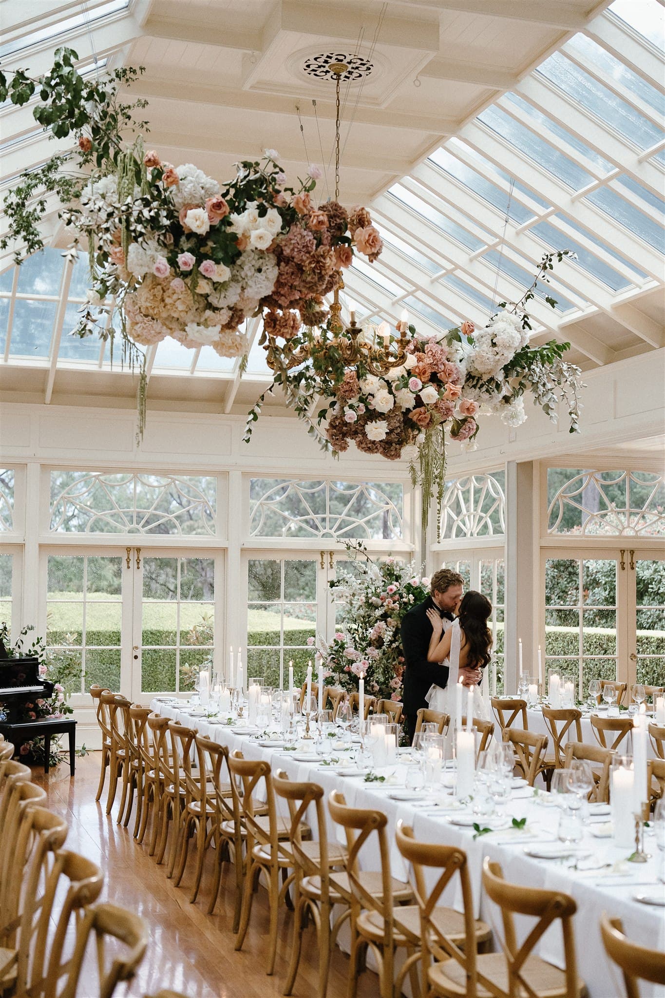 A couple shares an intimate moment in a beautifully decorated glass-roofed hall, surrounded by long tables adorned with white linens, candles, and floral centerpieces. Overhead, large hanging floral arrangements add to the romantic atmosphere.