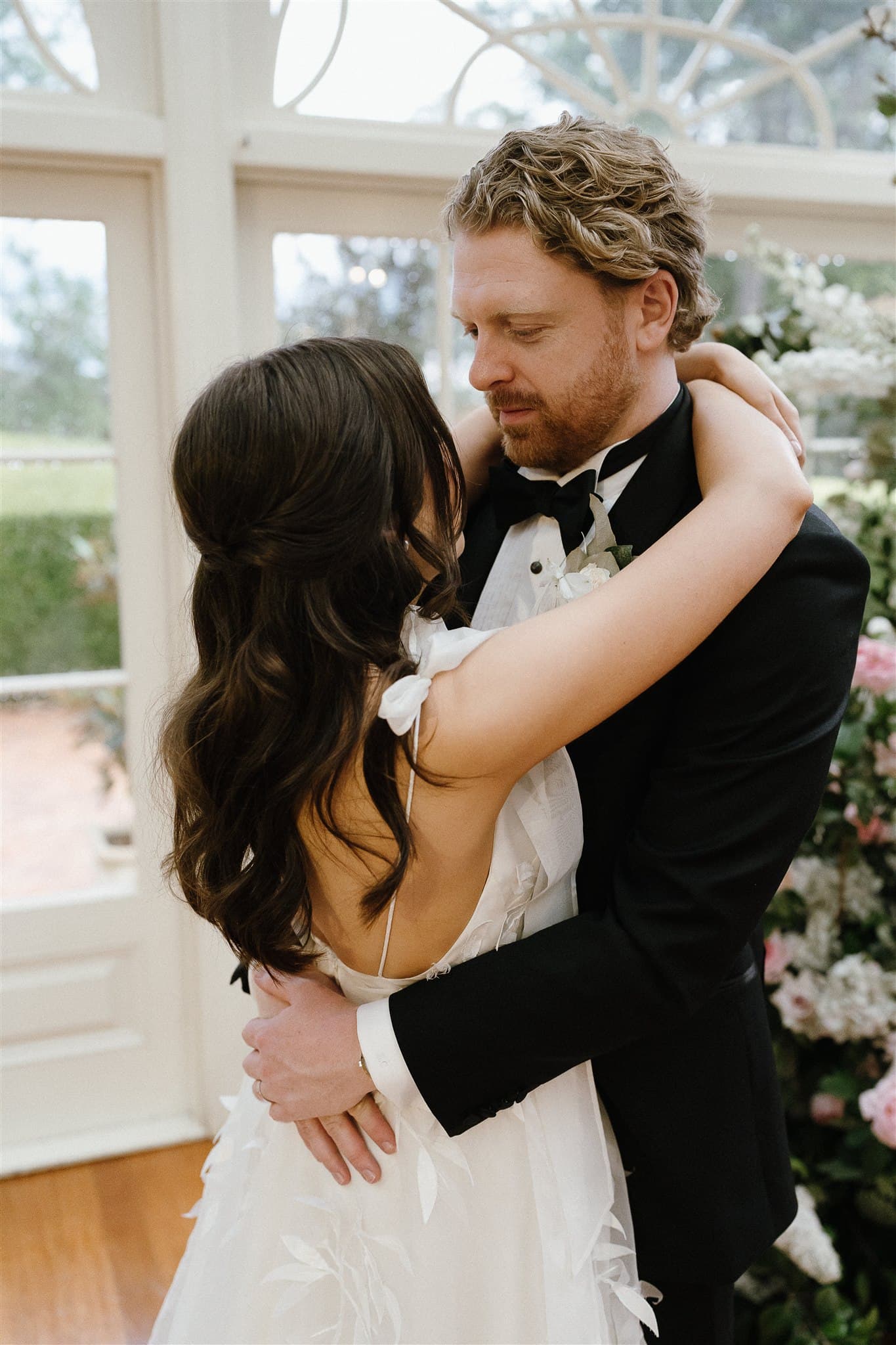 A couple dressed in wedding attire embrace each other lovingly in front of a window. The bride is wearing a white dress with her back to the camera, while the groom, in a black suit, gazes down at her. Flower decorations fill the background, creating a romantic scene.