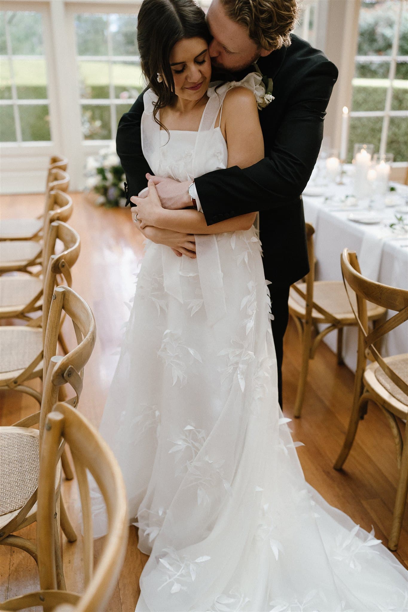 A couple dressed in wedding attire embraces in a warmly lit room with wooden chairs and a long, elegantly set table in the background. The bride, in a white gown adorned with floral appliqués, smiles as the groom hugs her from behind.