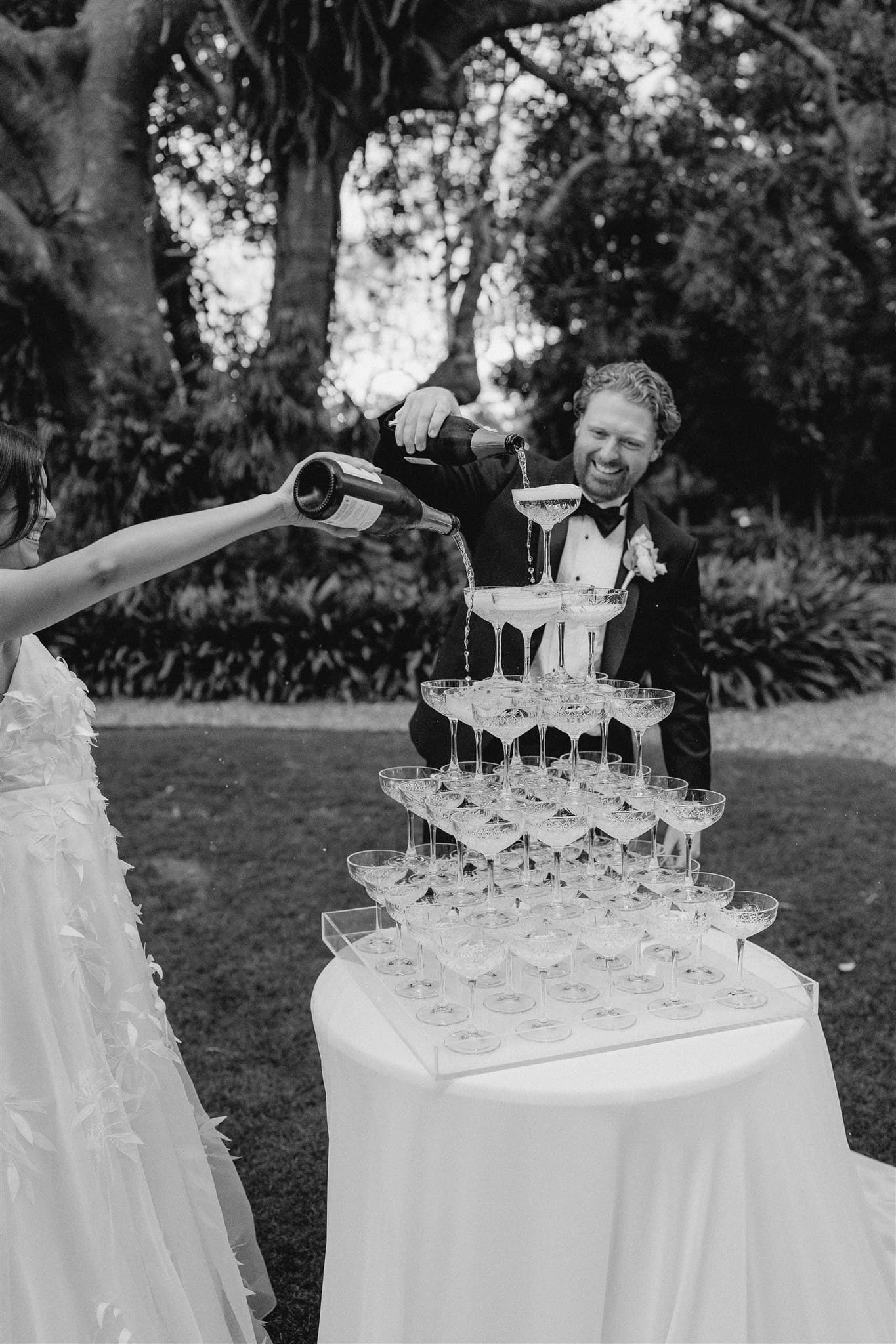 A person in a wedding dress pours champagne into the top glasses of a champagne tower on a white tablecloth outside. A person in a black tuxedo stands beside the table, smiling and watching the champagne cascade down the glasses. Trees and greenery are in the background.