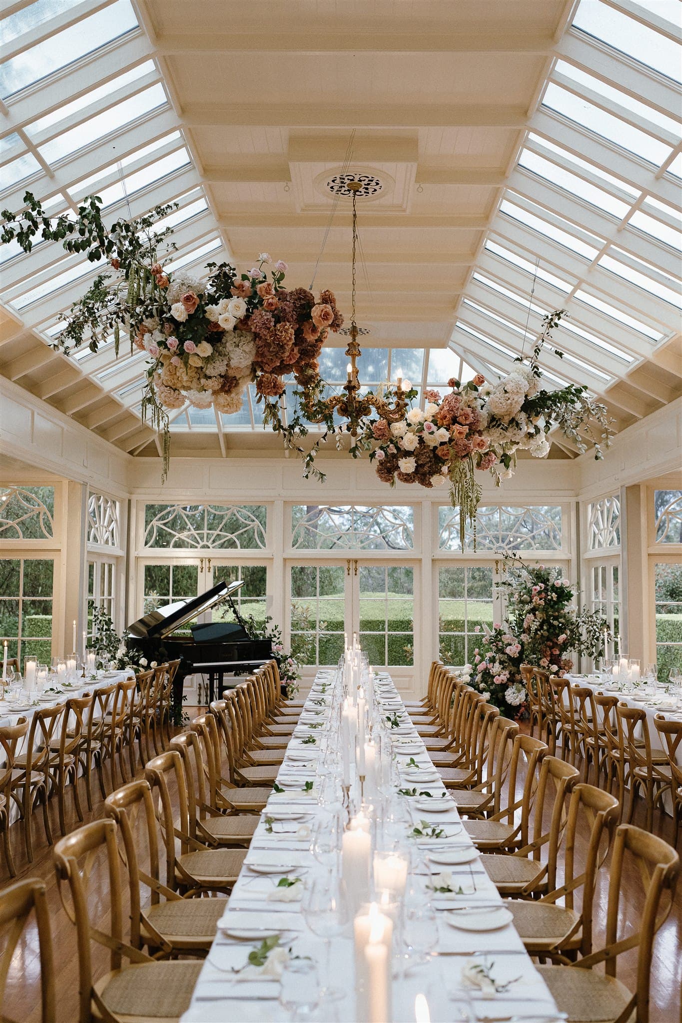 A beautifully decorated conservatory features a long dining table adorned with white linens, floral arrangements, and lit candles. Wooden chairs line either side of the table. Large floral chandeliers hang from the glass ceiling, and greenery is visible through tall windows.