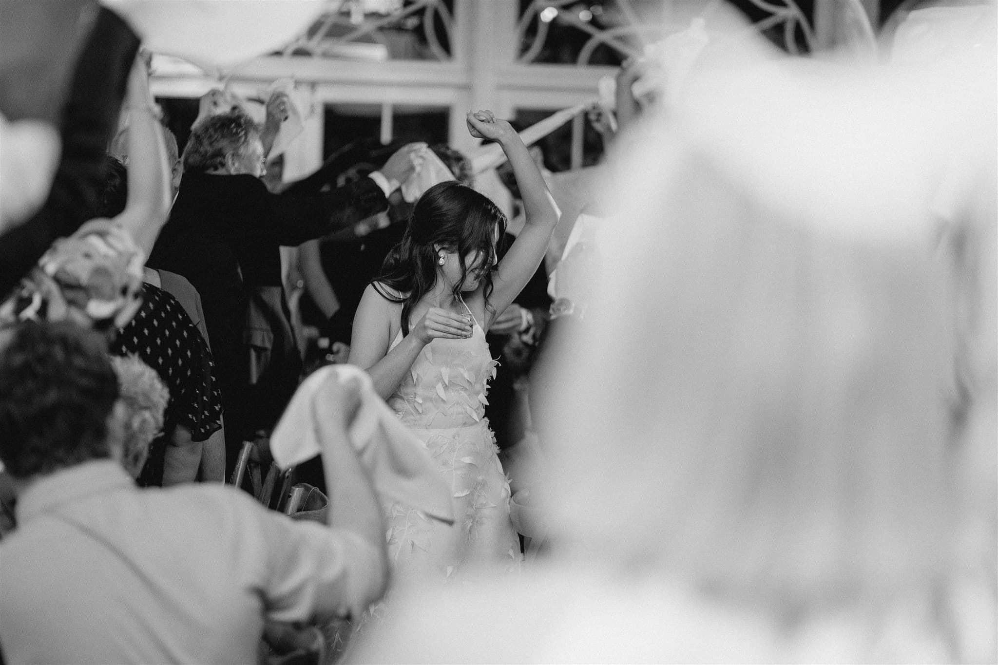 A black and white photo captures a festive scene where a young girl in a dress is energetically dancing among a crowd of people. The crowd is holding fabric or cloth, adding to the lively atmosphere. The lighting and expressions suggest a joyful celebration or event.