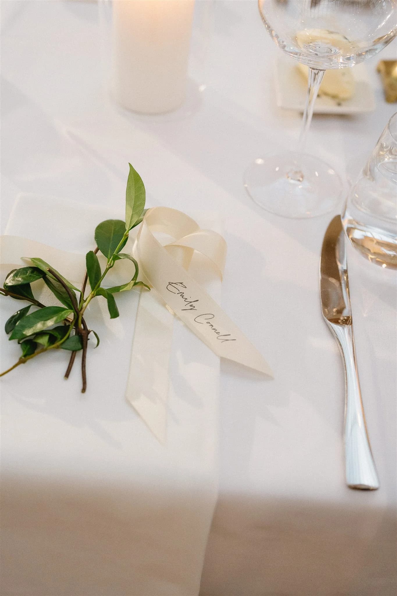 A place setting features a white ribbon with calligraphy text, greenery, a folded white napkin, and a candle on a white tablecloth. Nearby are a wine glass, a fork, and a knife. The ribbon is marked with the name "Emily Carroll.