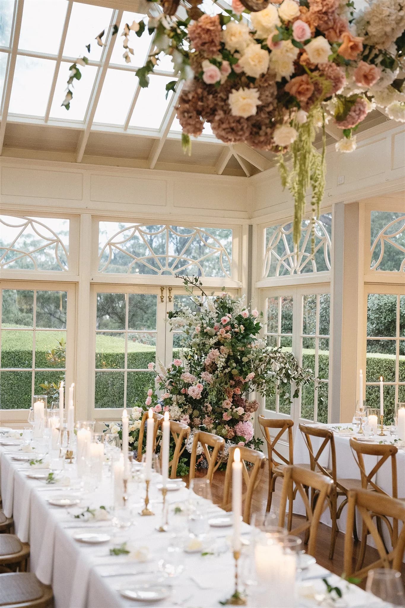 A beautifully decorated wedding reception room with floor-to-ceiling windows. Long tables are adorned with white cloths, numerous lit white candles, and elegant floral arrangements. A large hanging floral arrangement is above the table, adding to the ambiance.