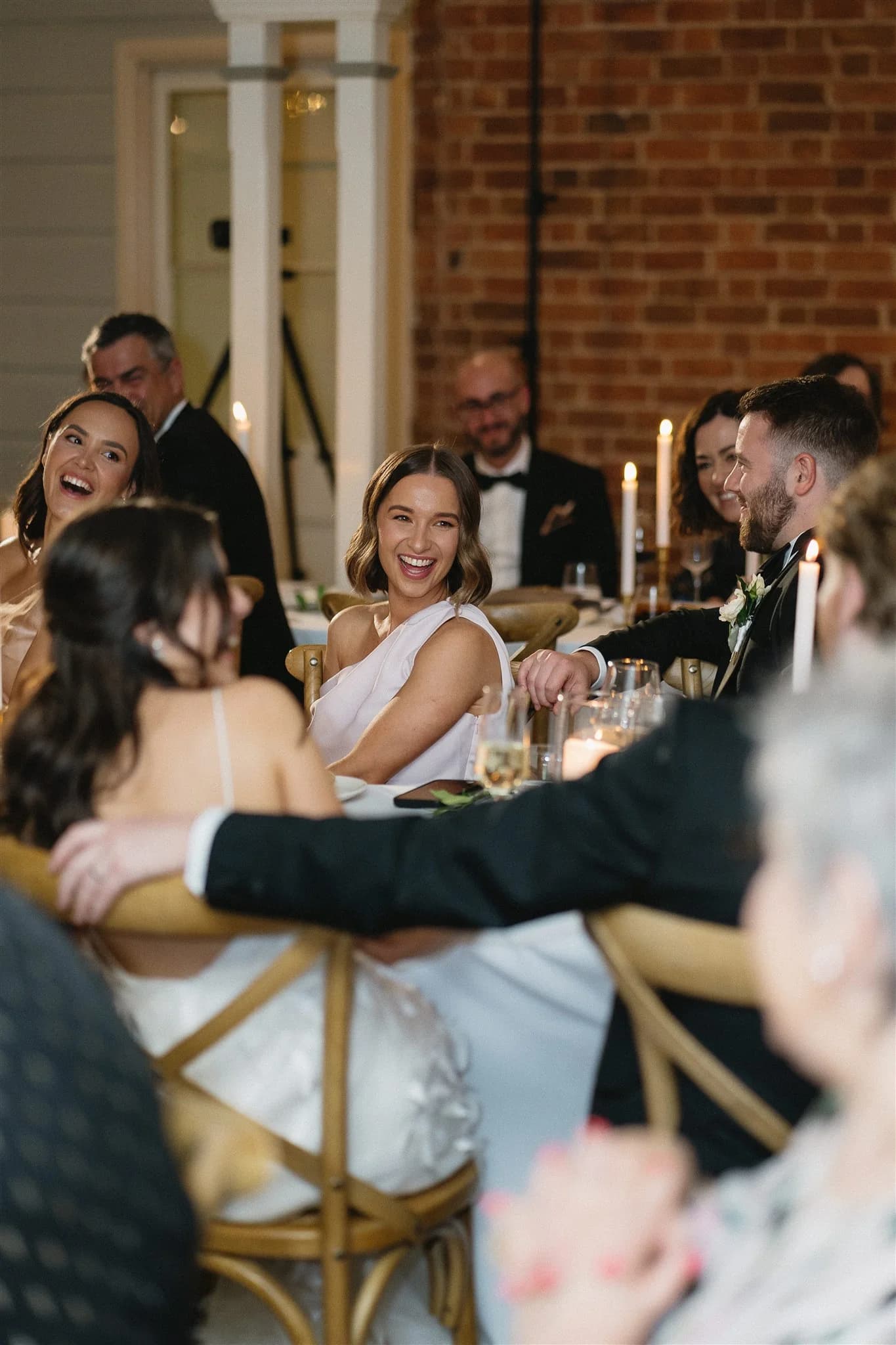 A group of people are seated together at a formal event, engaged in conversation and laughter. The atmosphere appears to be cheerful and celebratory, with candles and elegantly set tables adding to the ambiance. Women in dresses and men in suits can be seen.