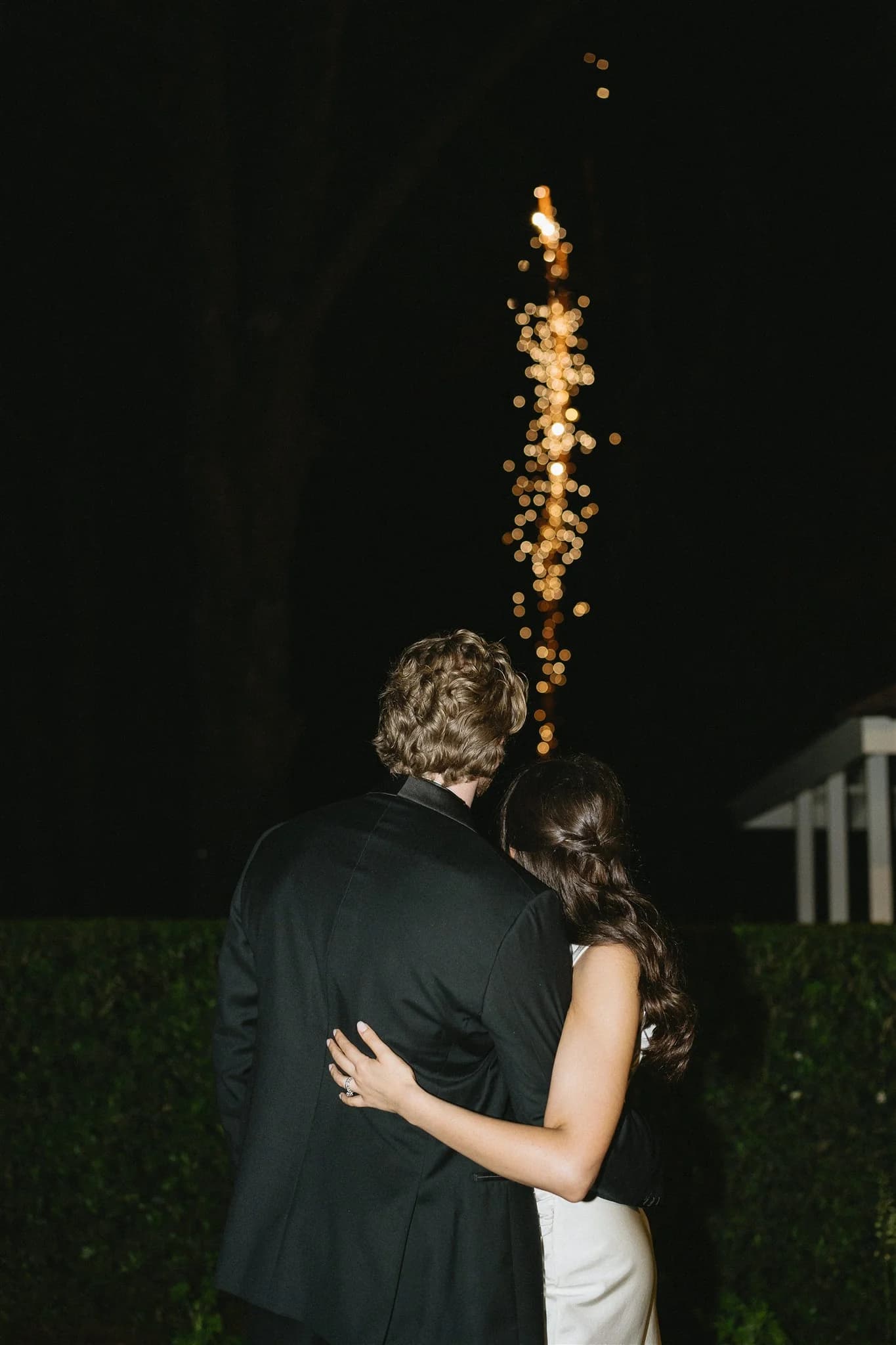 A couple dressed in formal attire stands closely together with their backs to the camera, watching golden fireworks light up the dark night sky. The woman has long, wavy brown hair, and the man is in a black suit with short, tousled hair.