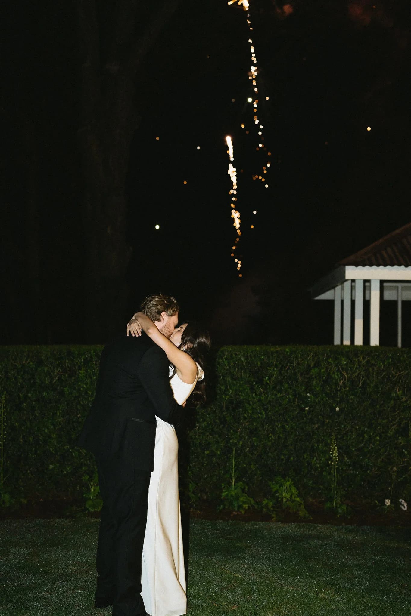 A couple embraces in front of a tall green hedge, illuminated by bright fireworks above them. The man wears a dark suit, and the woman is in a white dress. It is nighttime, and the scene is romantic, celebrating a special moment.