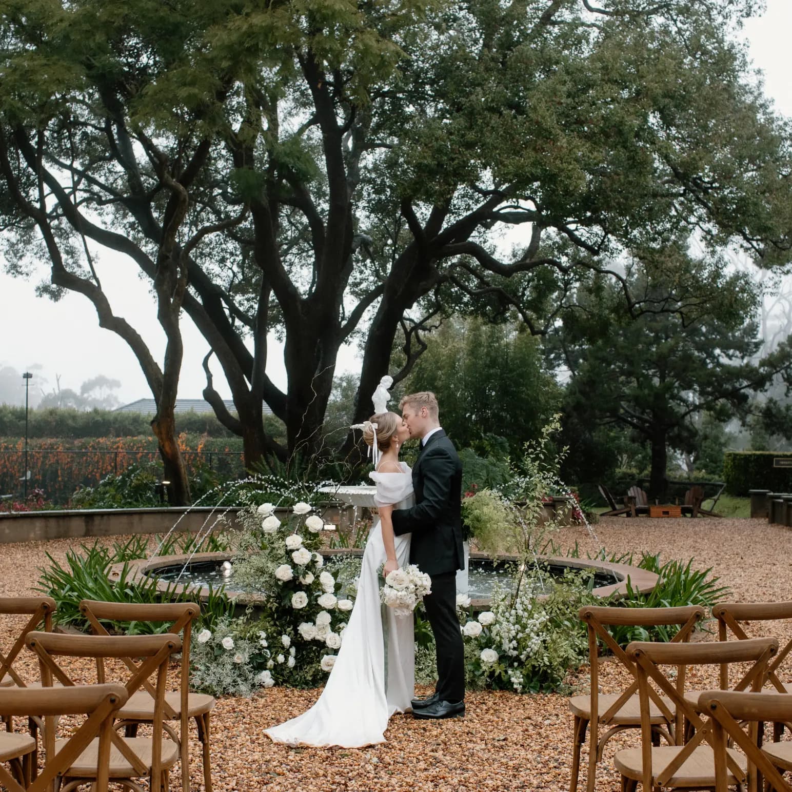 A couple in wedding attire stands in front of a lush garden with a large tree and a circular arrangement of white flowers and greenery. They are sharing a kiss, surrounded by wooden chairs arranged for guests on a gravel pathway. Hill and trees are visible in the misty background.