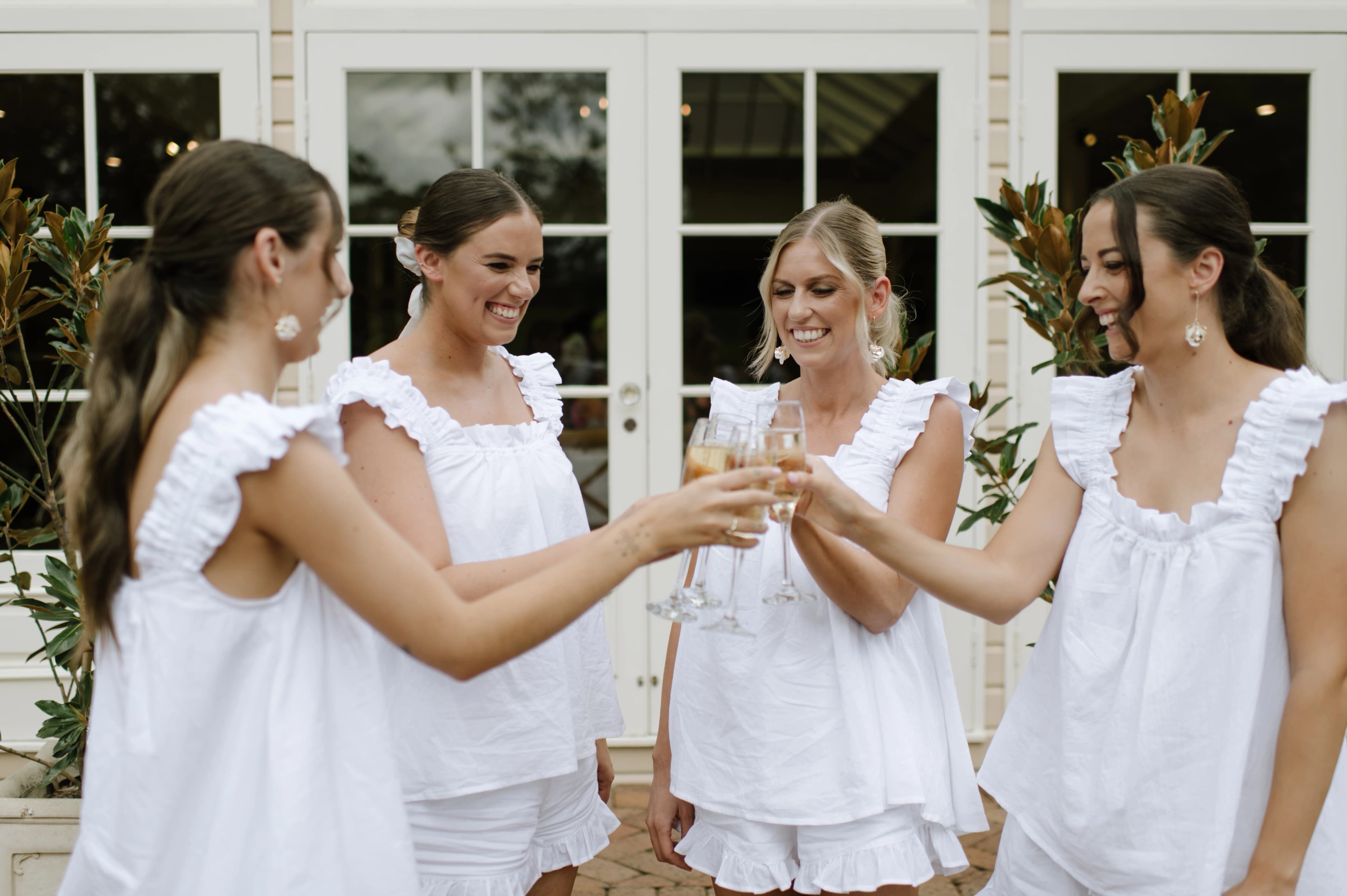 four women in elegant white summer pajama sets raising champagne glasses in a toast, celebrating together.