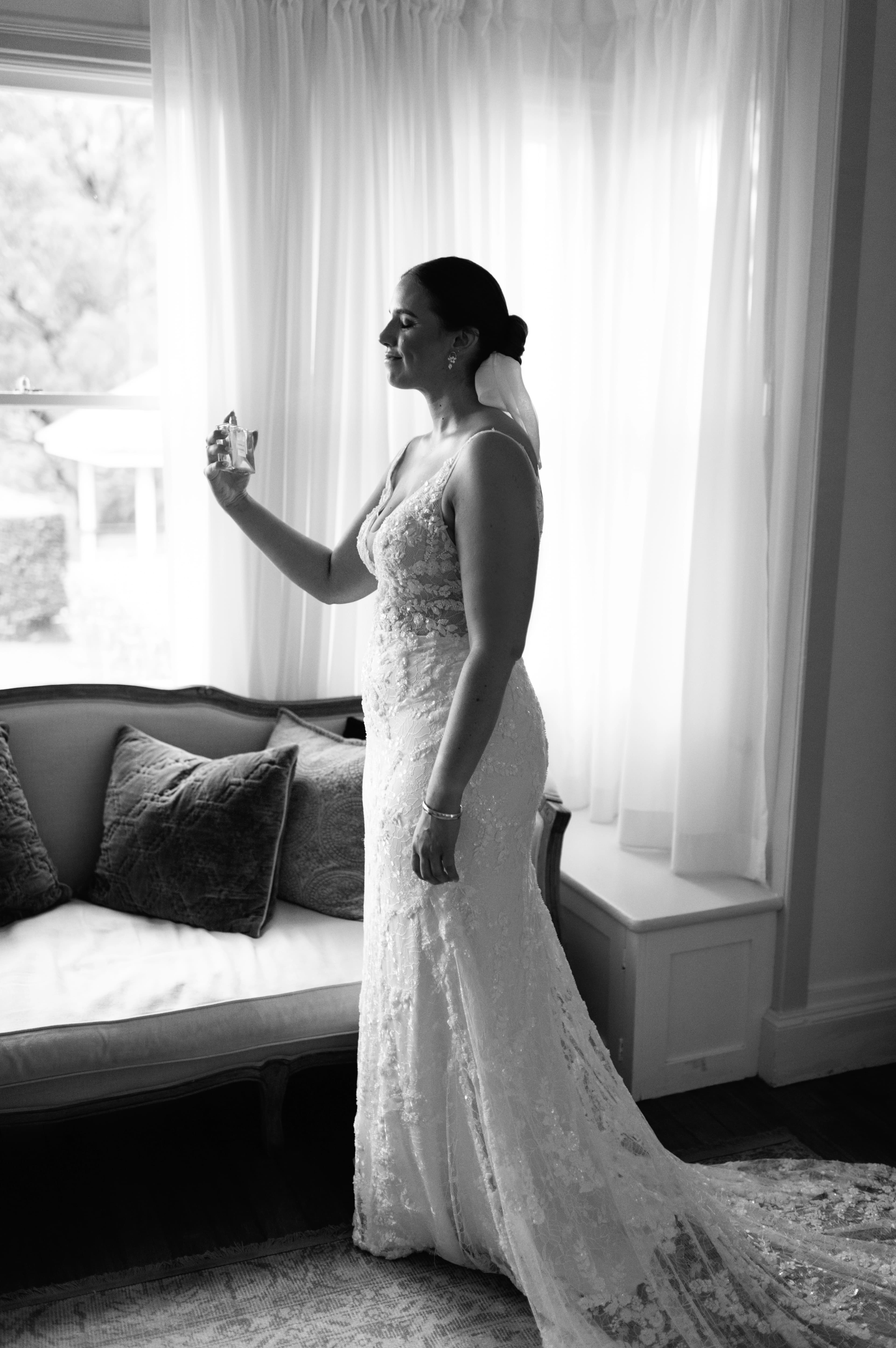 A bride, dressed in her wedding gown, applying perfume while standing by a window