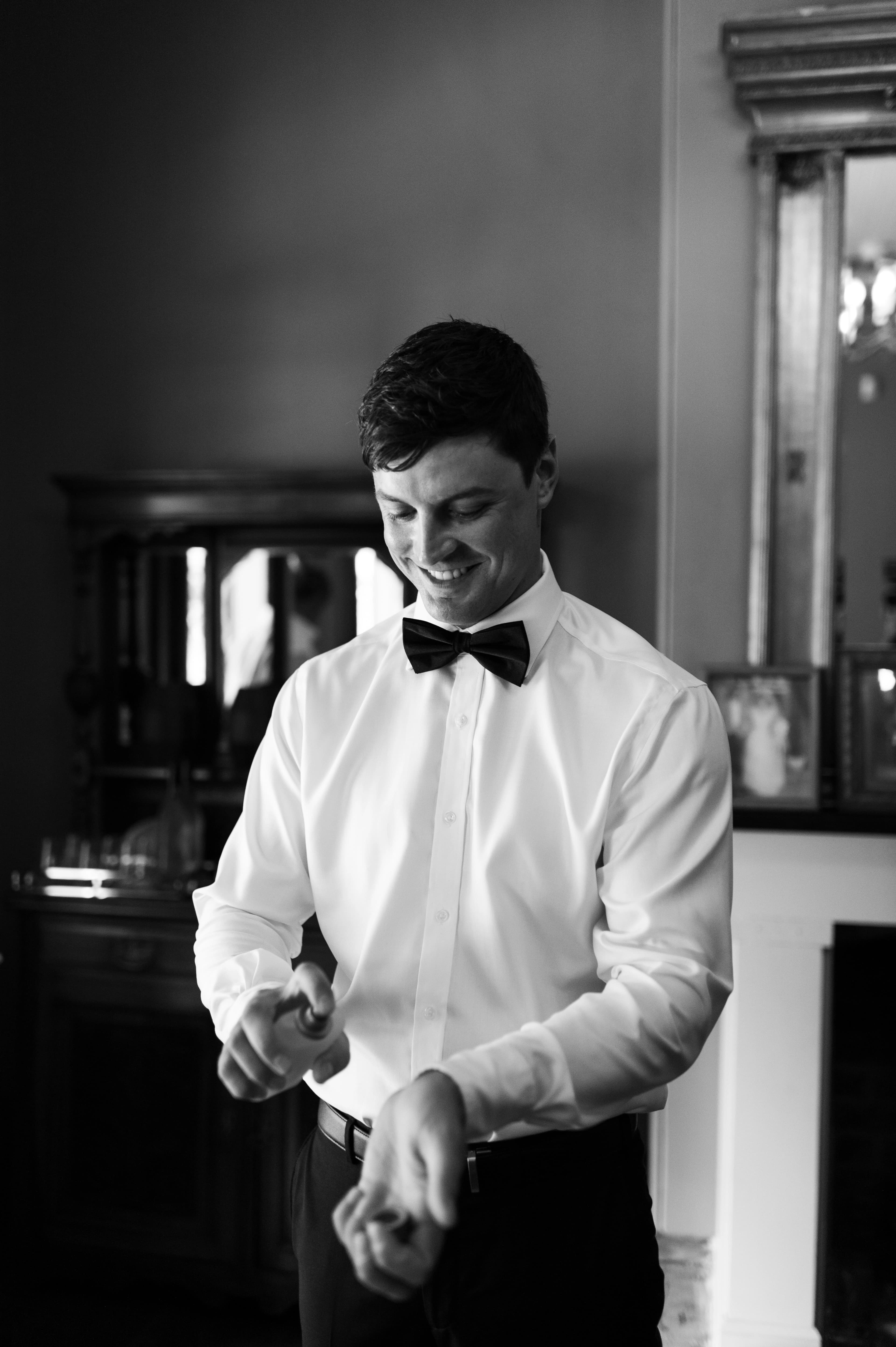 A man dressed in a bow tie and shirt, smiling warmly while spraying cologne on his wrist.