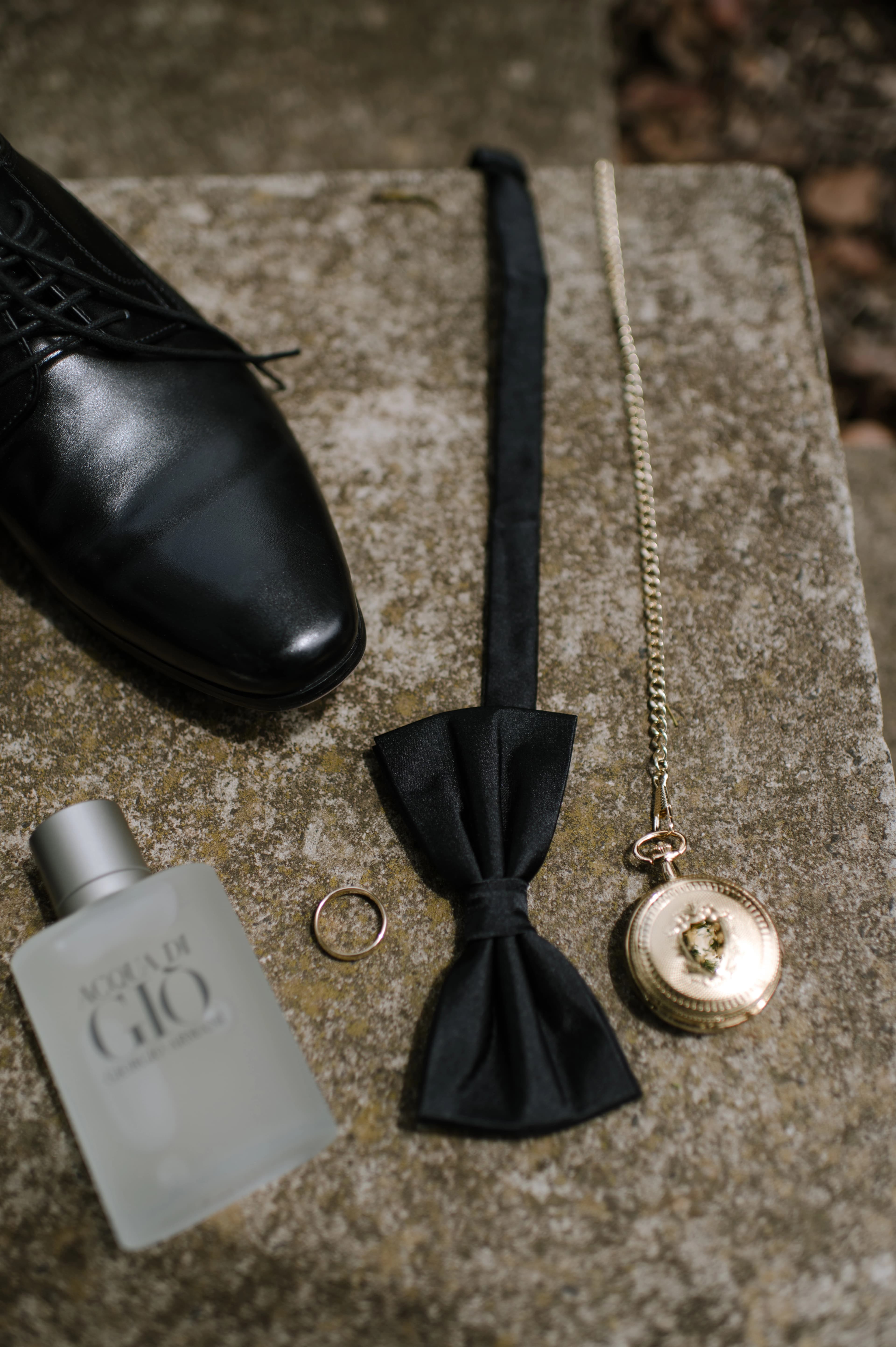 A black shoe, a gold ring, a perfume and a vintage pocket watch arranged on a neutral background for a wedding.