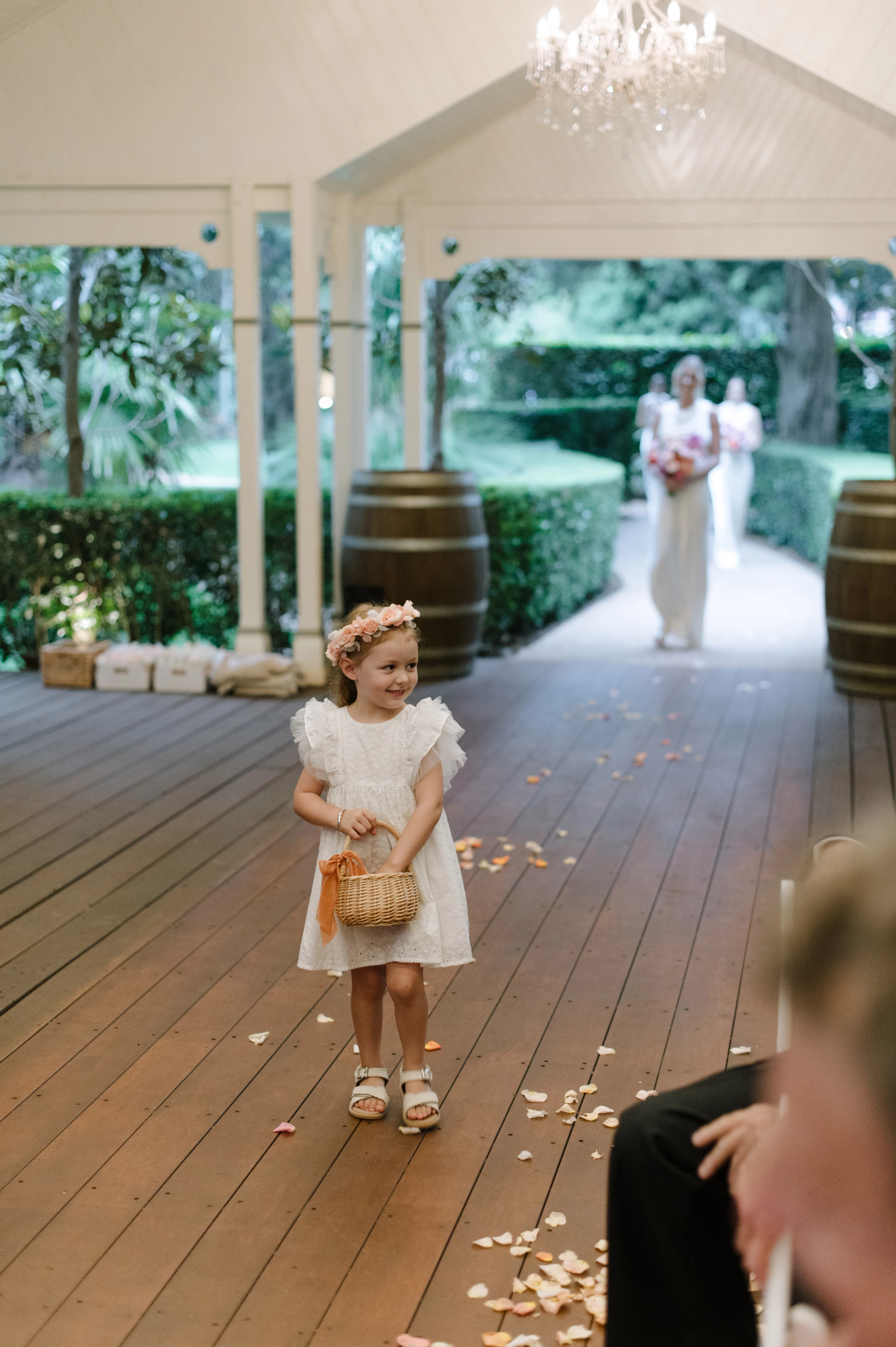 A young flower girl in a white dress smiles while holding a basket of petals at a wedding ceremony